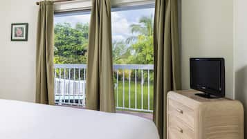 A hotel room with a bed, a television on a wooden dresser, and a balcony with a white railing and greenery.