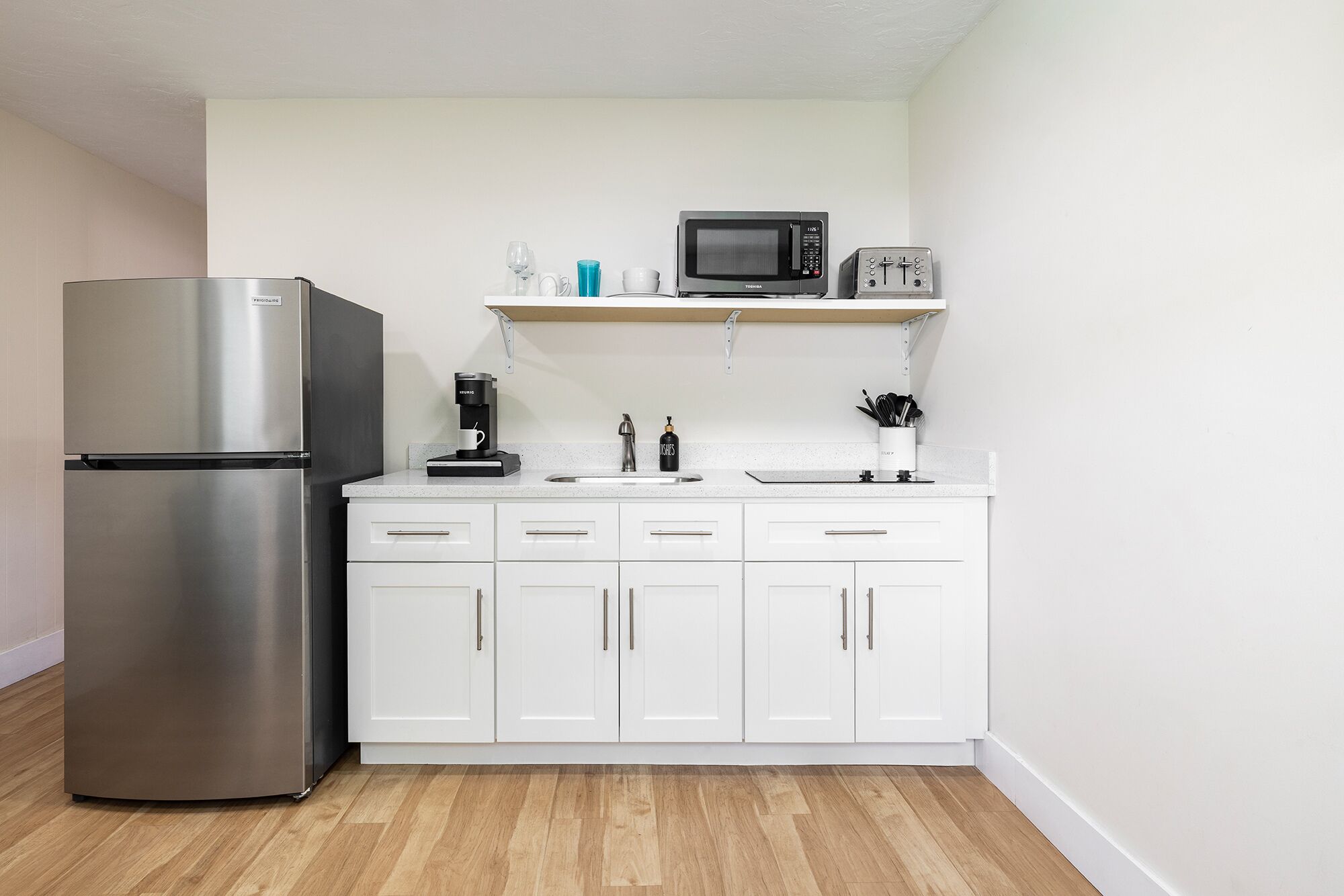 Una cocina compacta con refrigerador de acero inoxidable, microondas, cafetera y armarios blancos.