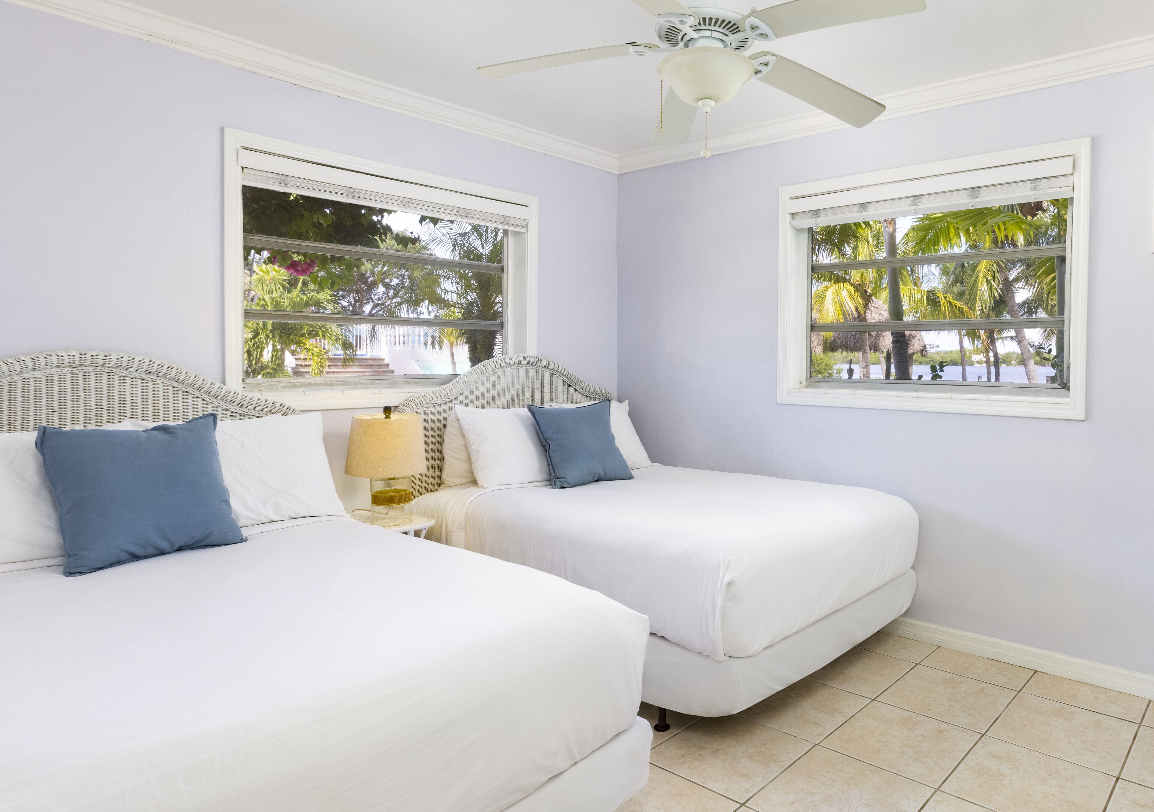 A room with two beds, a ceiling fan, and windows with views of palm trees.