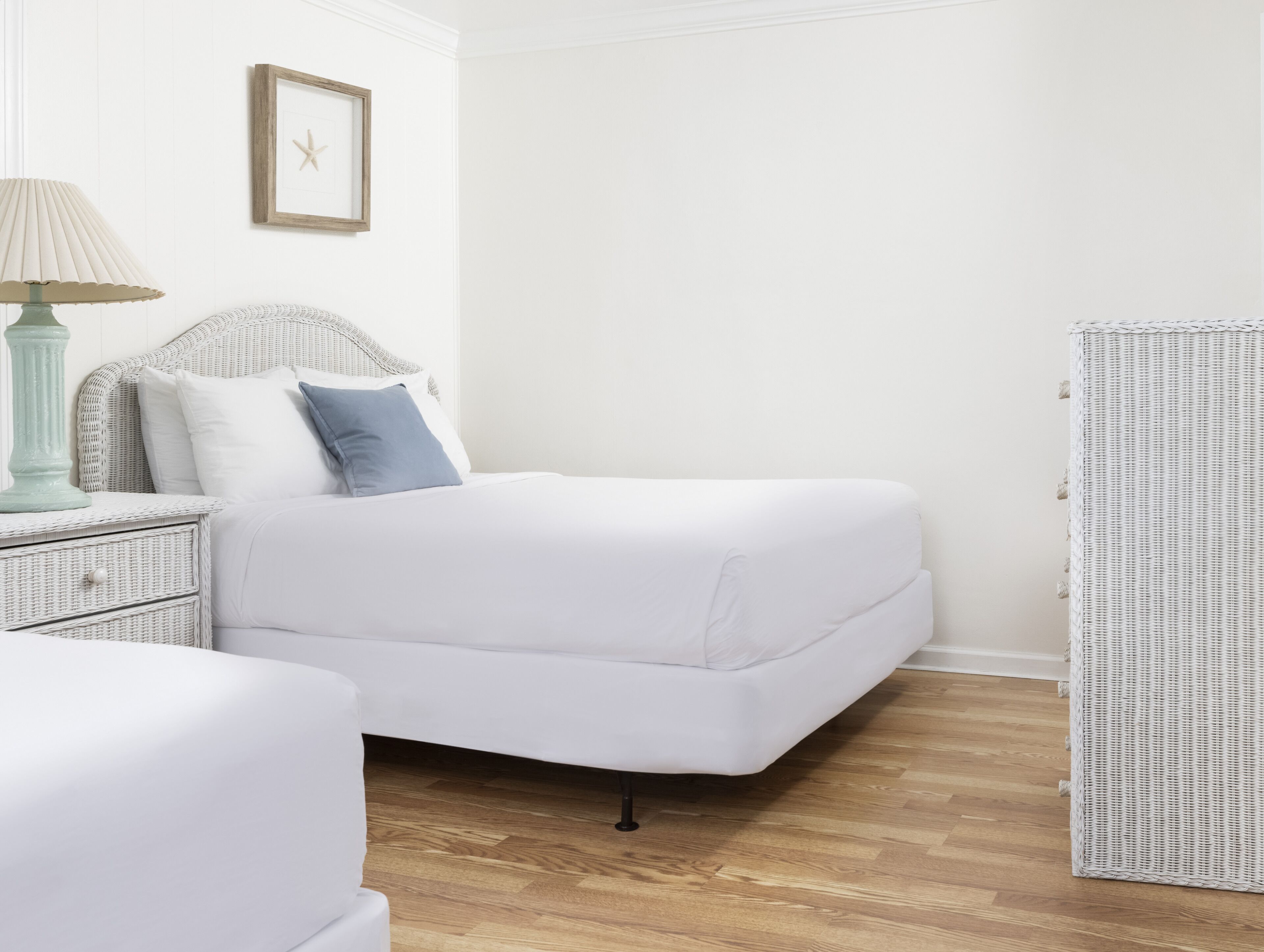 A white bed with a headboard, a bedside table with a lamp, and a wicker chair.