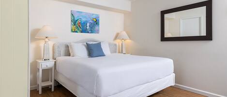 A neatly made bed with white linens, two bedside lamps, a painting of tropical birds, and a mirror on the wall.
