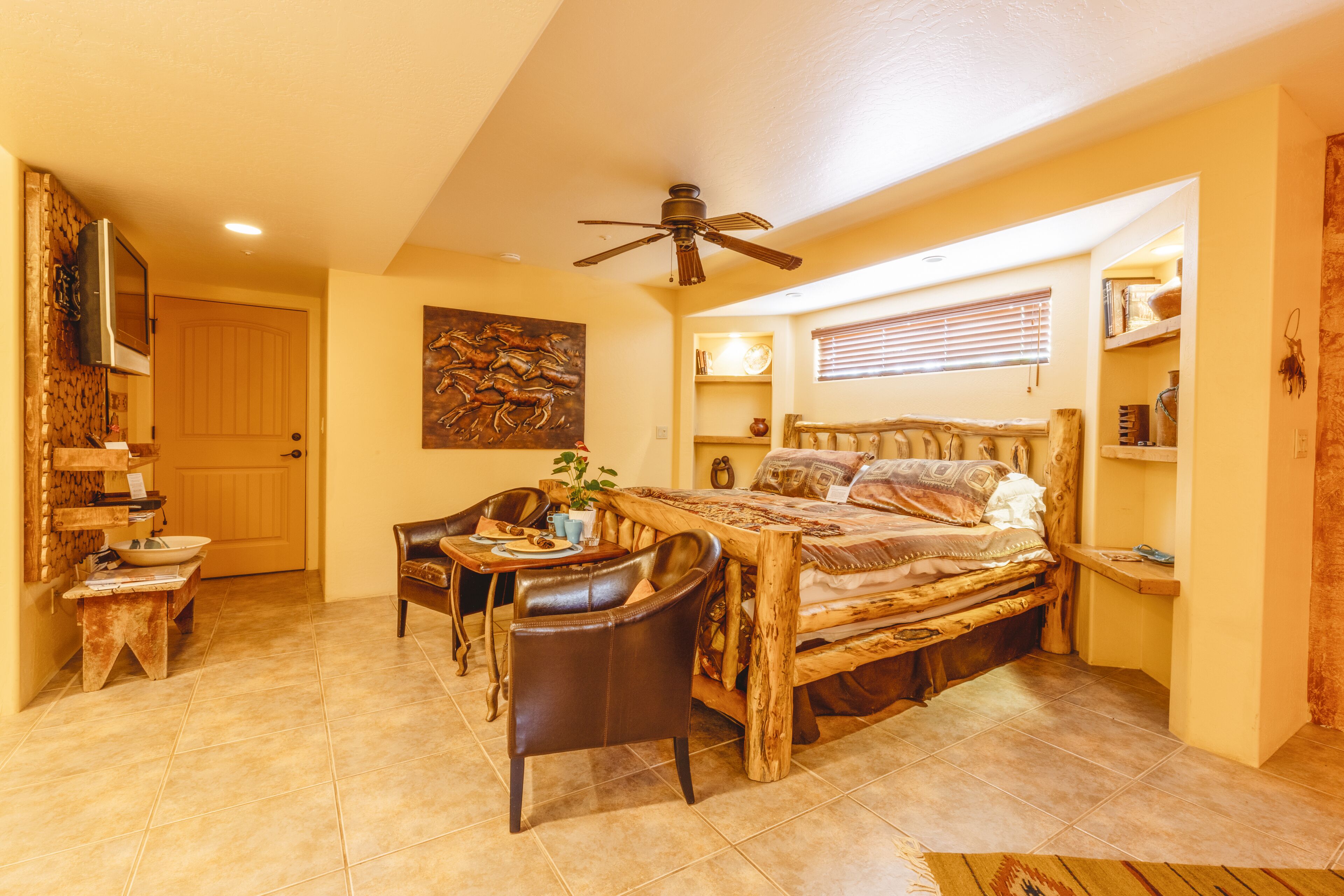 A cozy bedroom with a wooden bed, a dining table with chairs, a ceiling fan, and a door leading to another room.