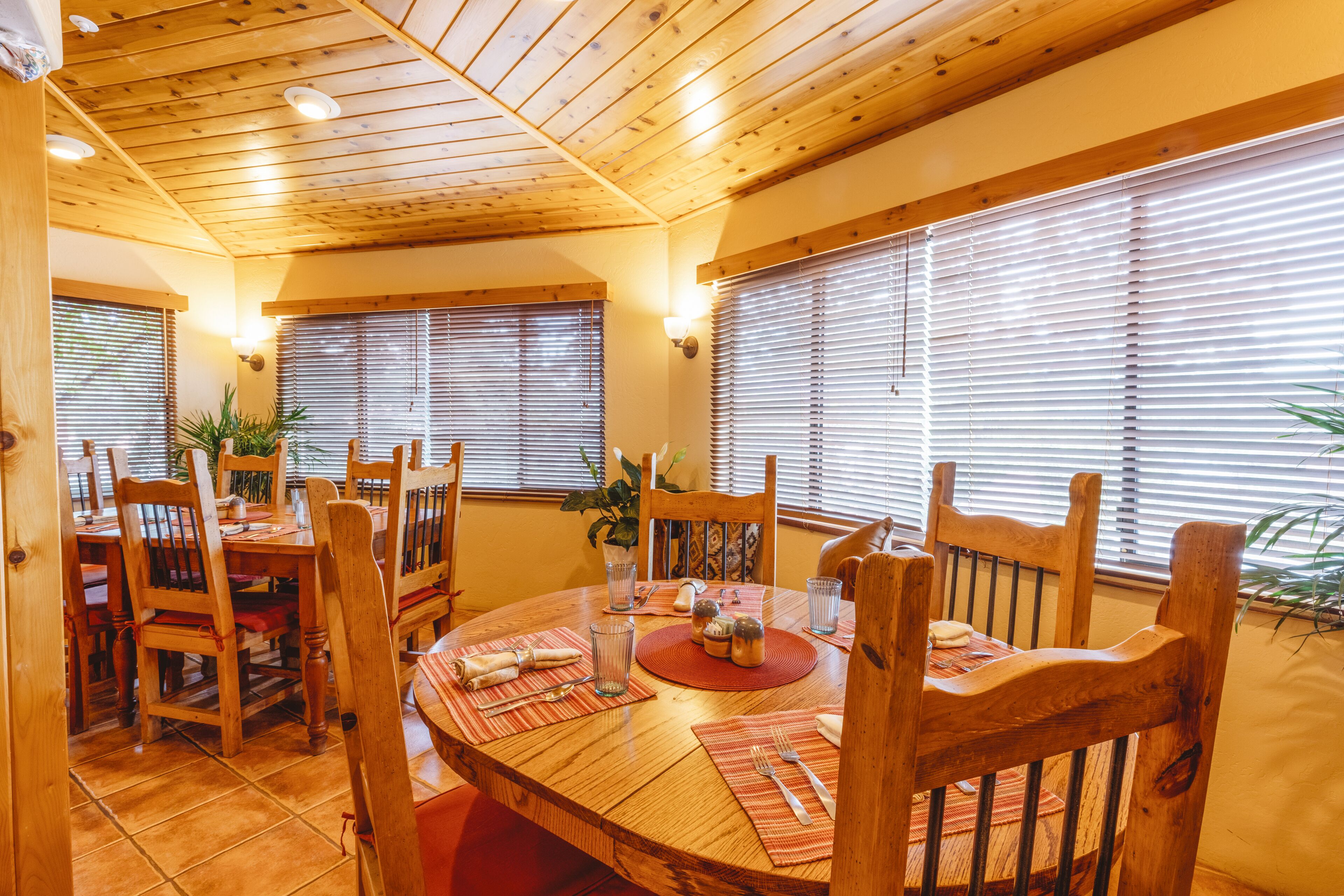 Ein Holzesstisch mit Stühlen, umgeben von Fenstern mit Jalousien, und eine Decke mit Holzbalken.