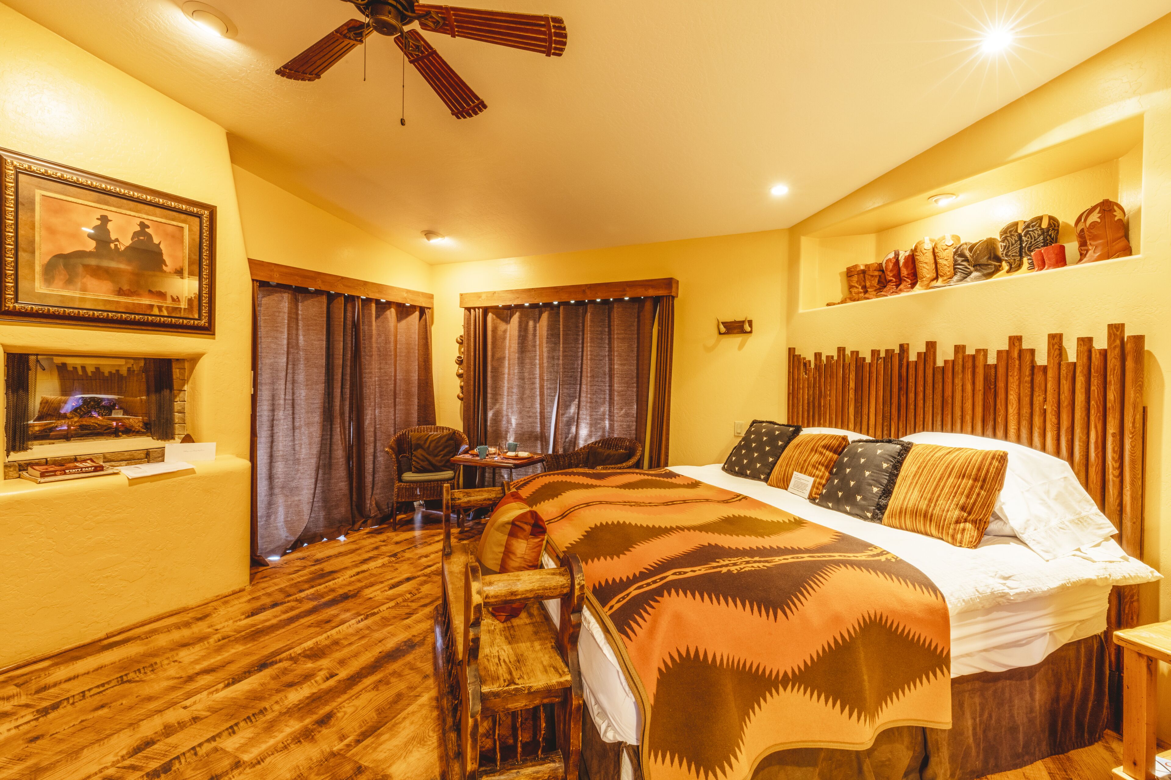 A hotel room with a wooden floor, a bed with patterned bedding, a ceiling fan, a mounted picture, a small table with a chair, and a shelf with boots.