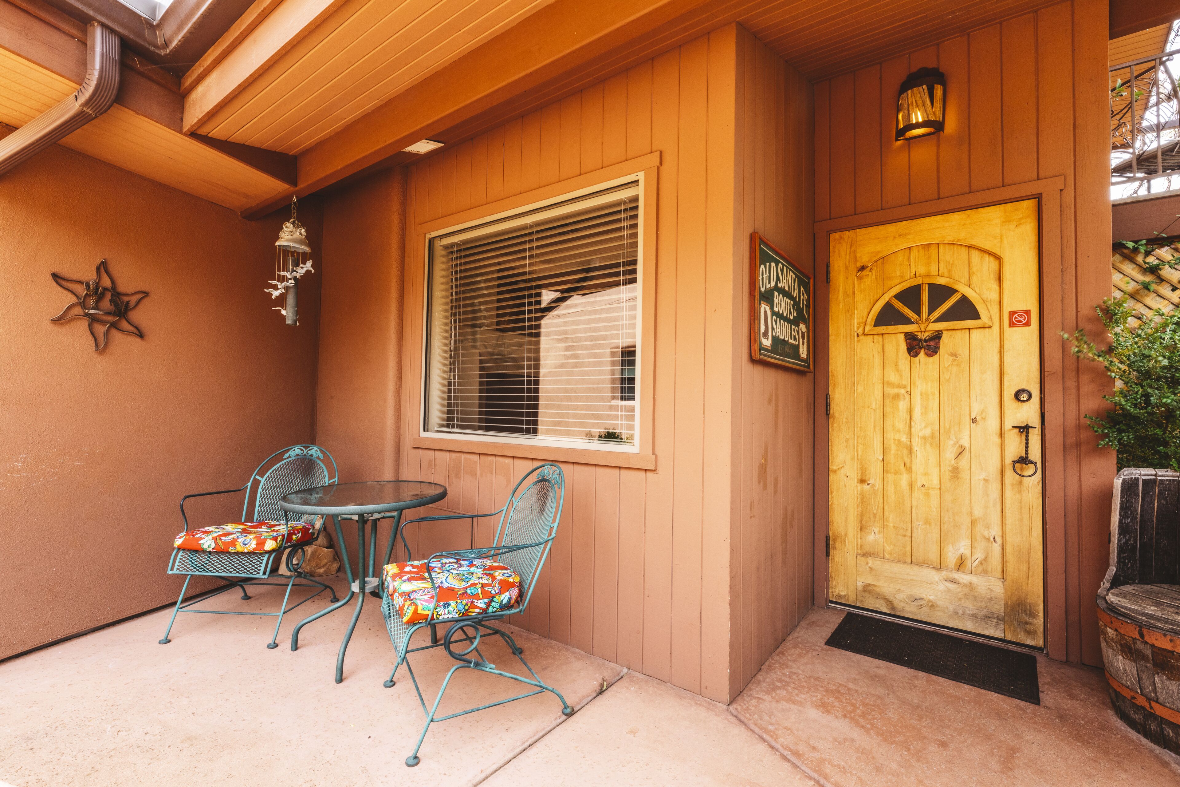 A wooden building with a wooden door, a window with blinds, and two metal chairs with colorful cushions.