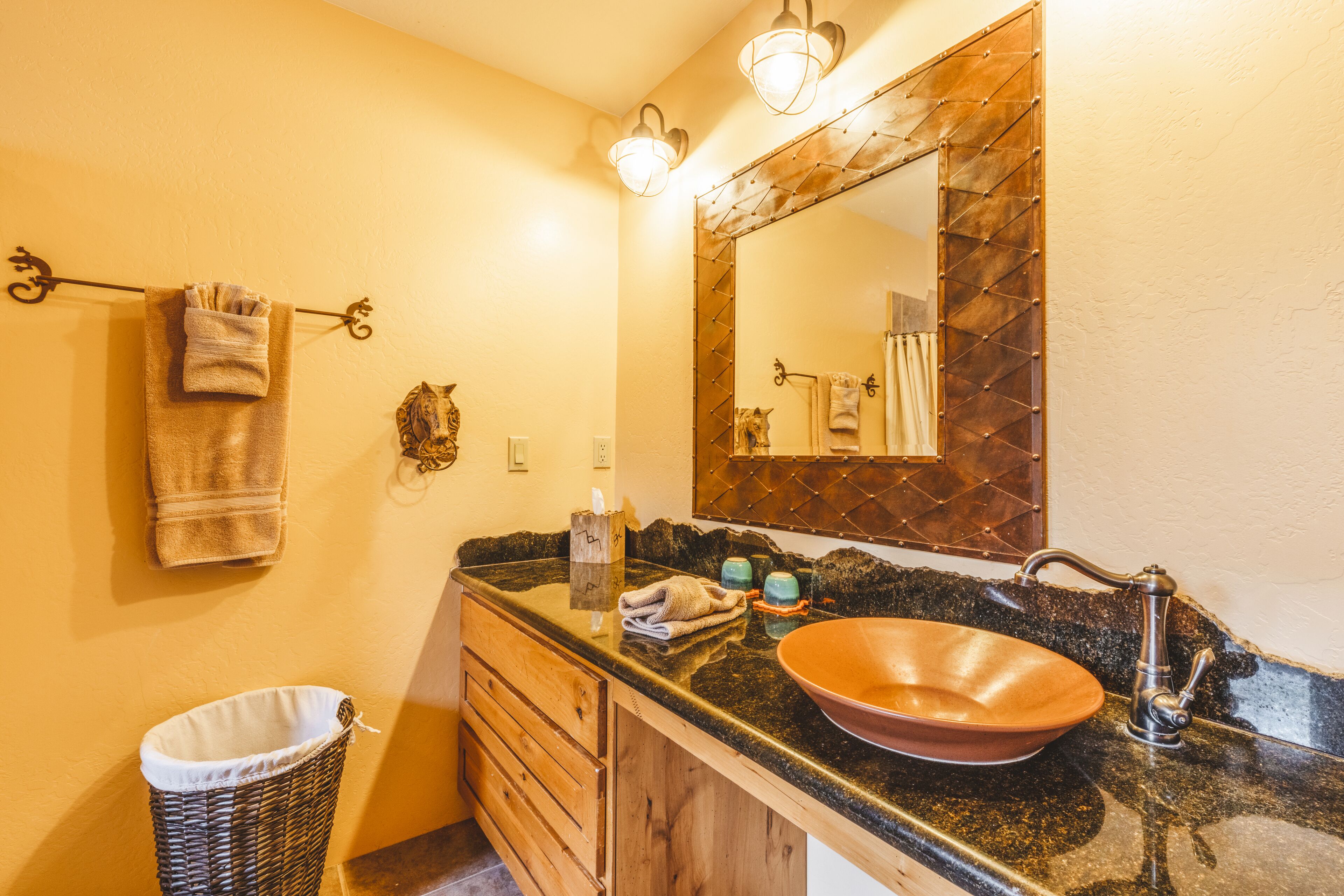 Un baño con encimera de granito negro, tocador de madera, un espejo grande con marco de diseño de rombos y una toalla colgada en un perchero.