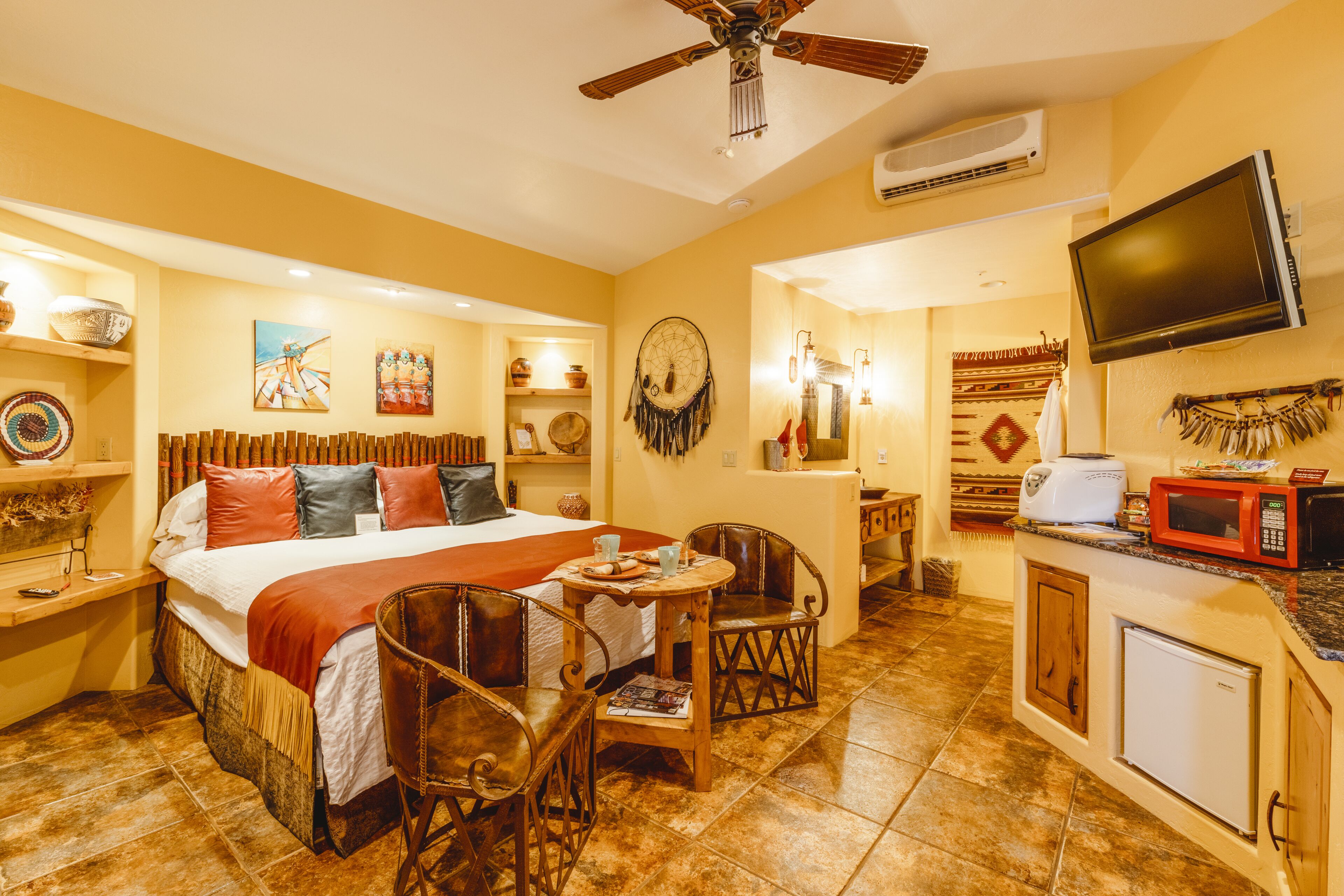 A hotel room with a bed, a dining table with chairs, a kitchenette, and a TV mounted on the wall.