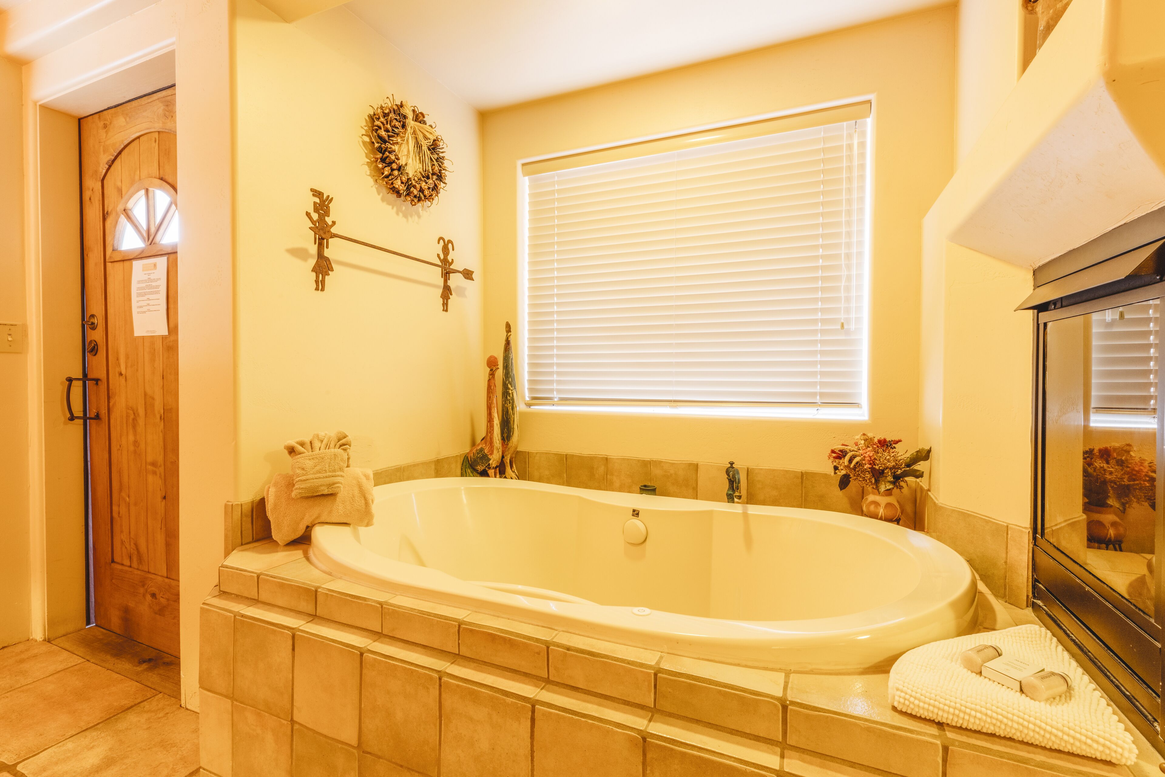 A bathroom with a large bathtub, a wooden door, a window with blinds, and a mirror.