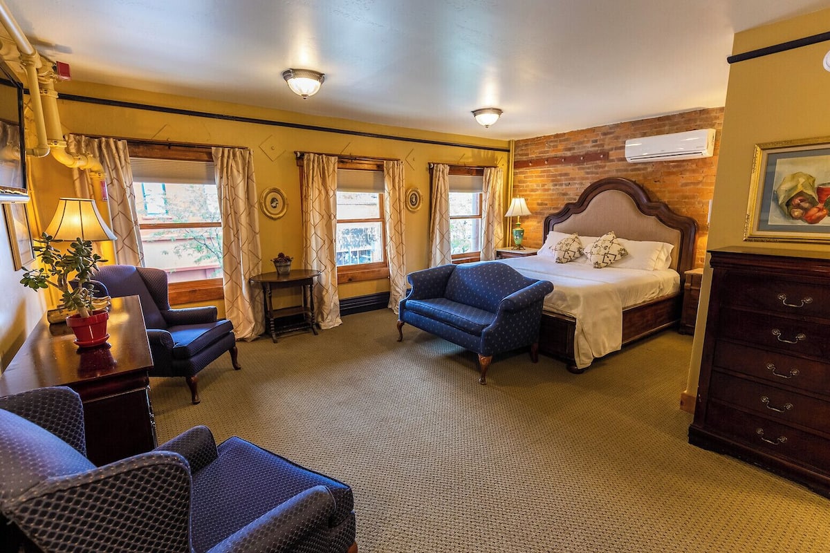 A hotel room with a bed, two armchairs, a small table, a dresser, and a window with curtains.