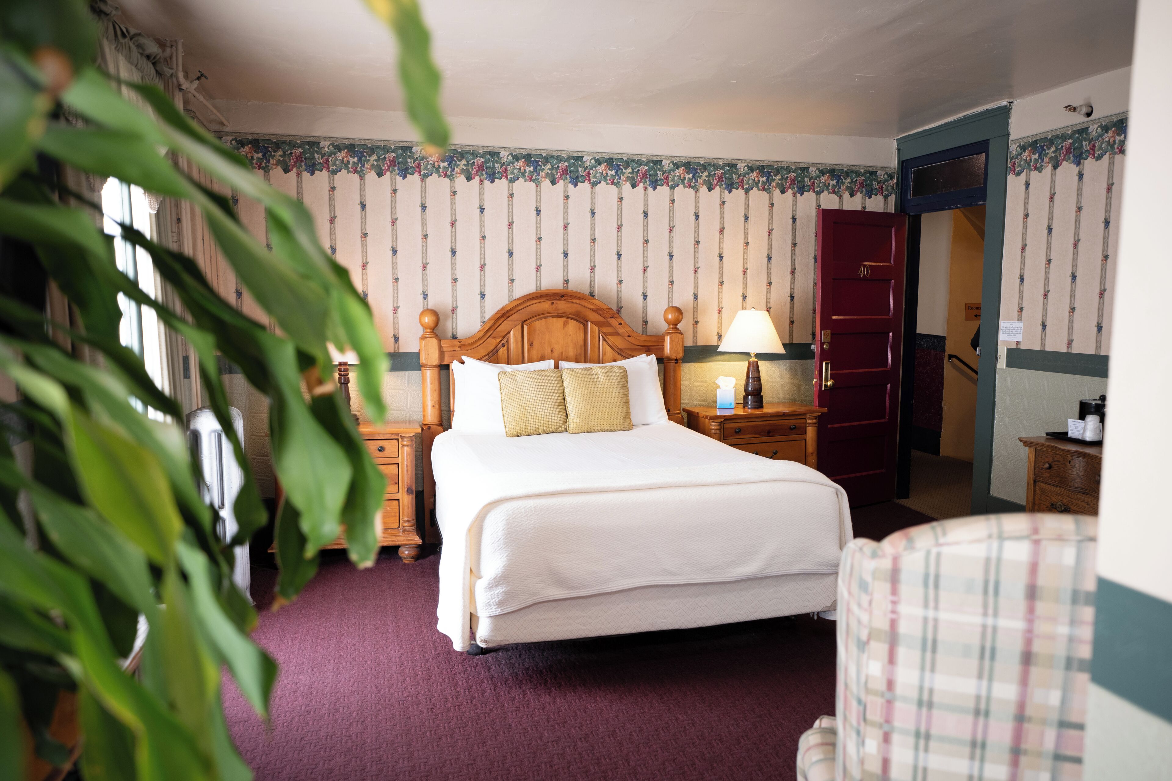 A hotel room with a large bed, wooden headboard, a nightstand with a lamp, a red door, and a plaid chair.