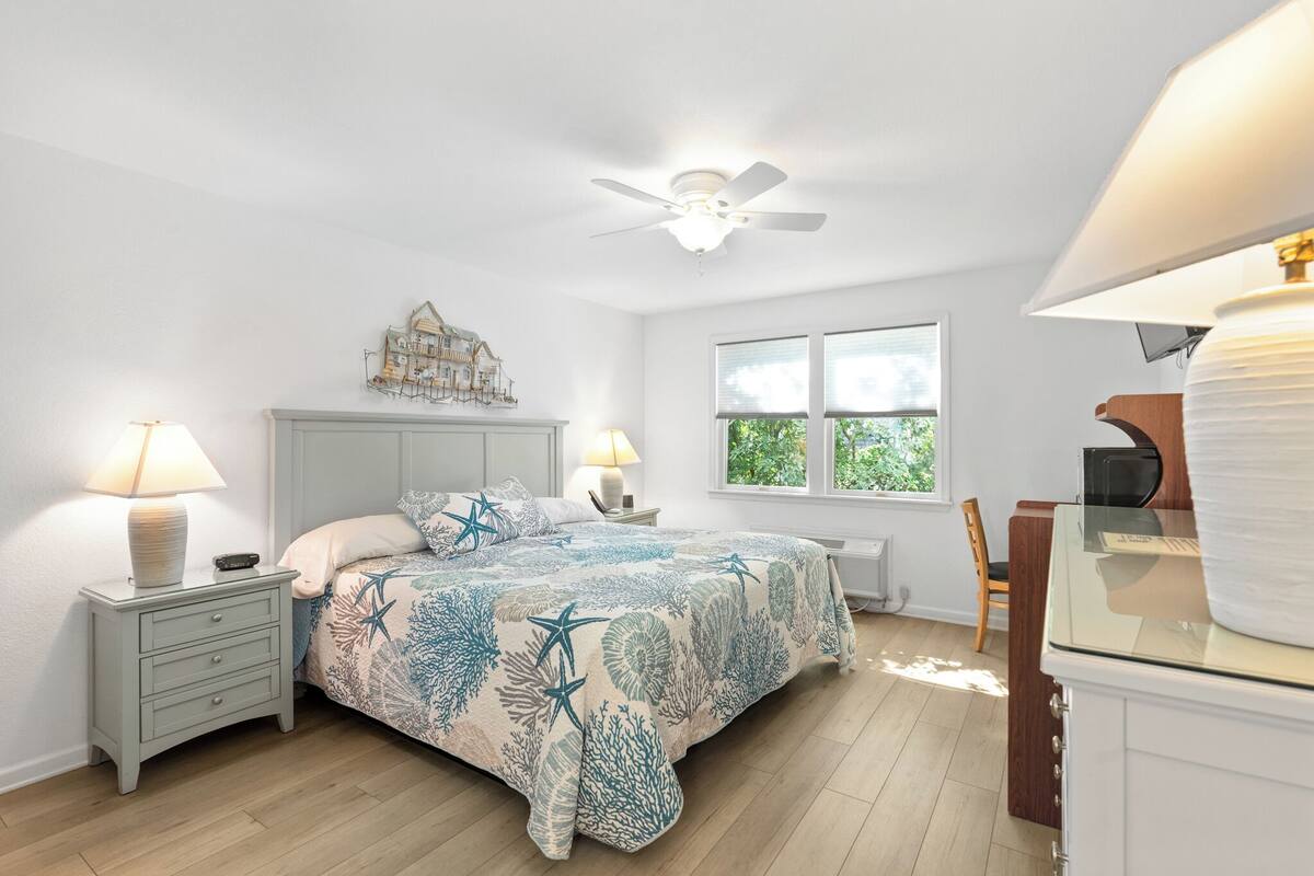 Ein Schlafzimmer mit einem Bett, Nachttischen, einem Deckenventilator, einem Fenster und einem Schreibtisch mit Lampe.