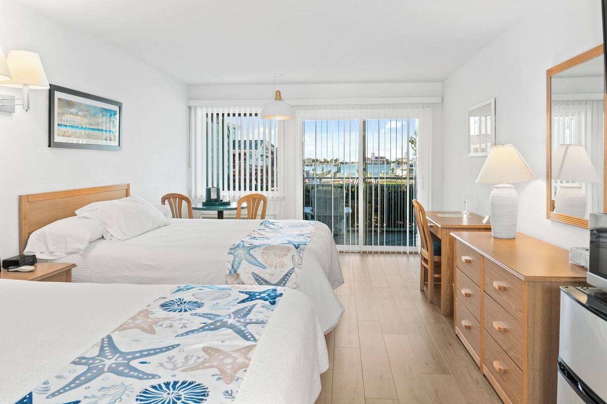 Ein Hotelzimmer mit zwei Betten, einem Schreibtisch, einem Stuhl, einem Schminktisch und einem Balkon mit Meerblick.