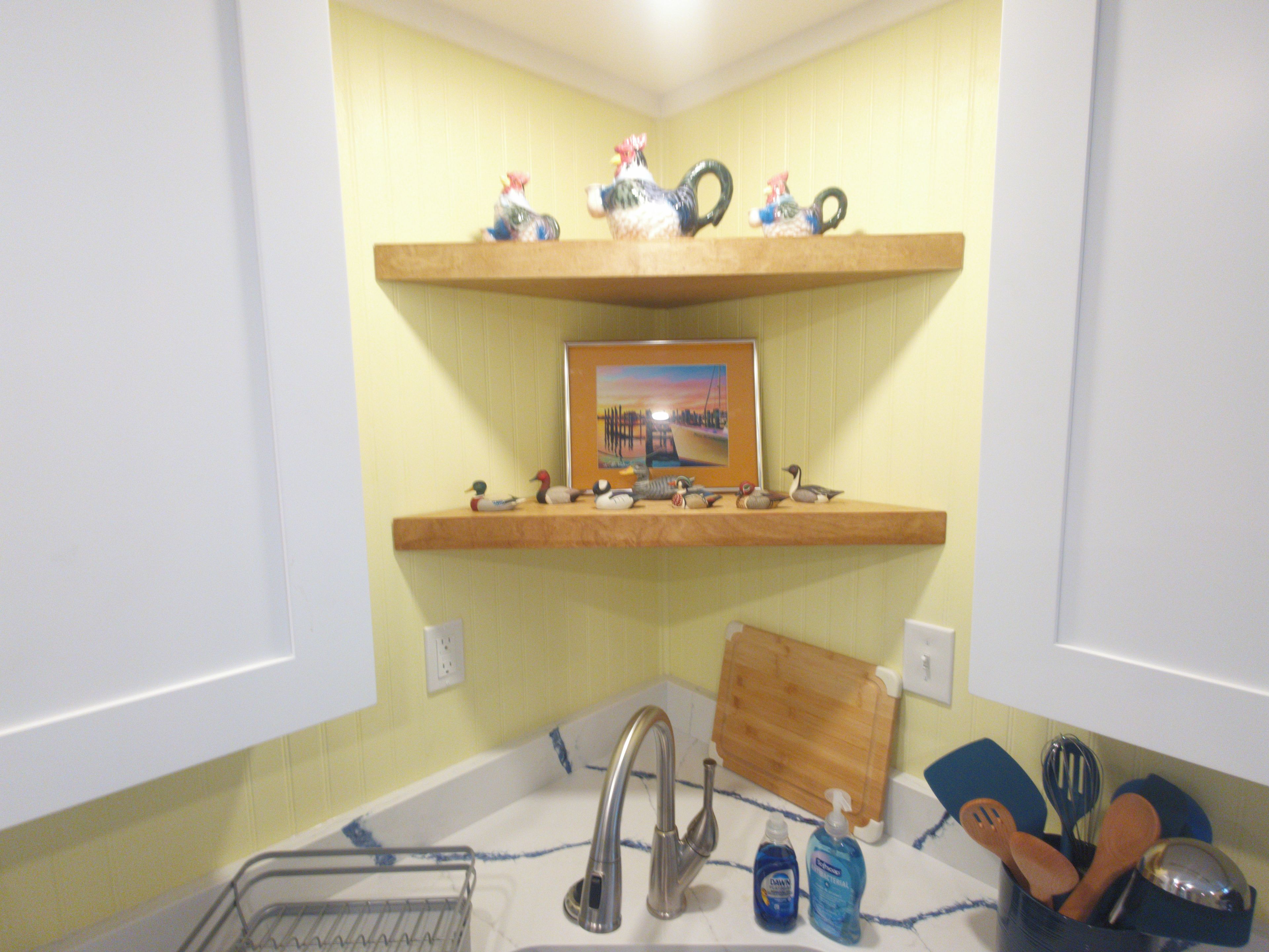 A bathroom with a wooden shelf holding decorative items and a framed picture, a sink with a faucet, and a cutting board on the countertop.