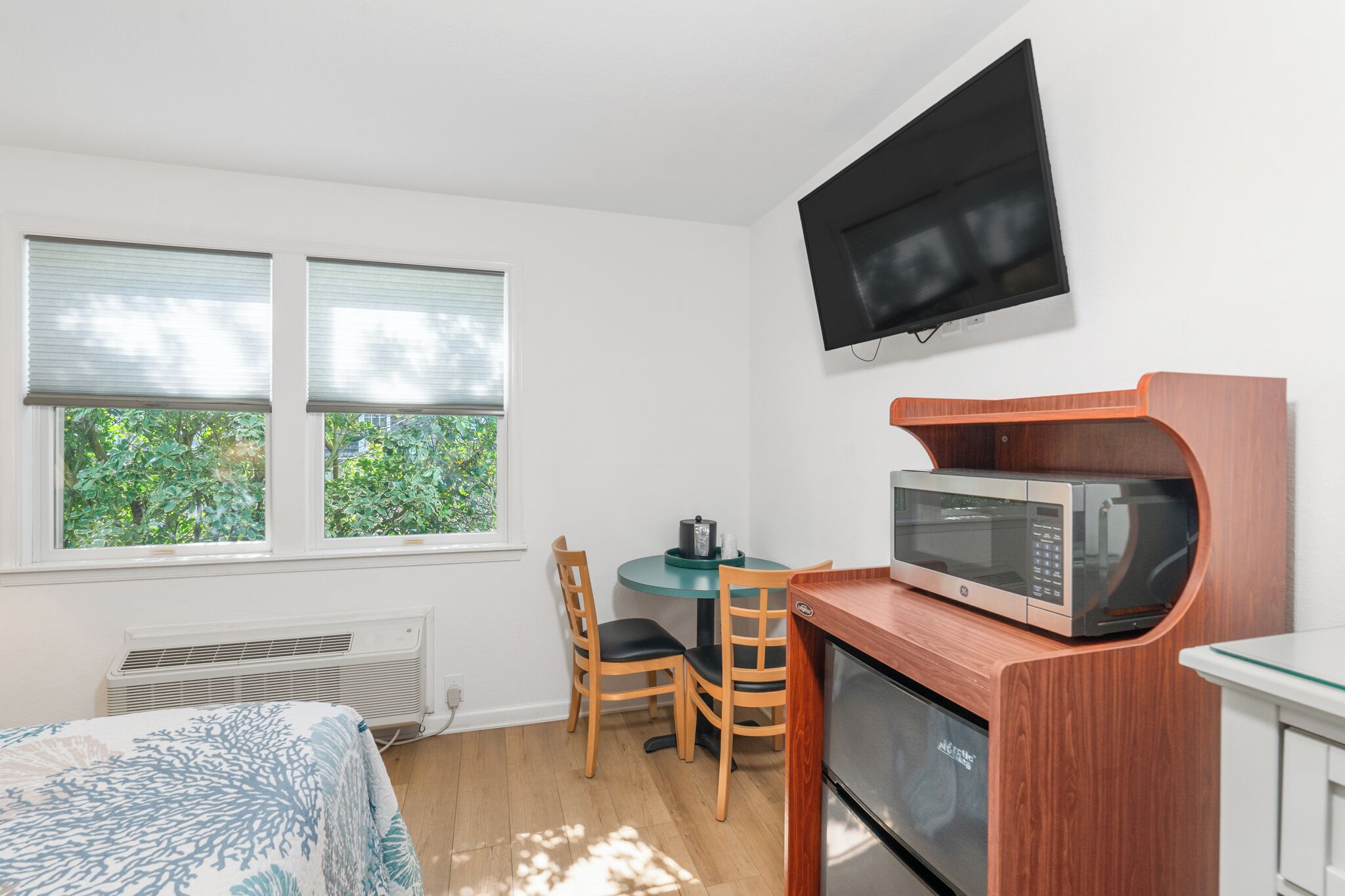 A compact room with a bed, a small dining table with chairs, a microwave, and a TV mounted on the wall.