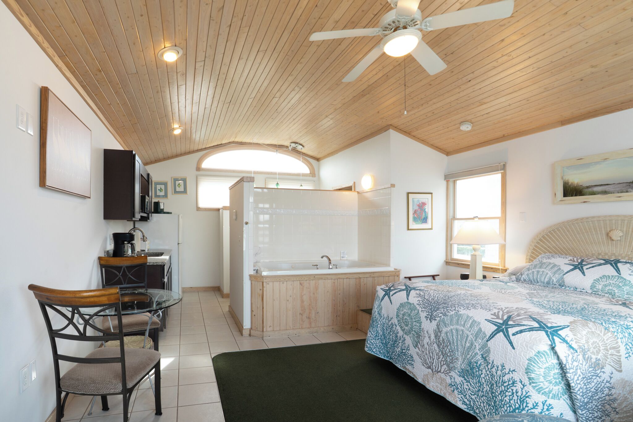 A room with a wooden ceiling, a bed with a starfish-themed bedspread, a small kitchenette, a bathroom with a bathtub, and a dining area with a glass table and chairs.