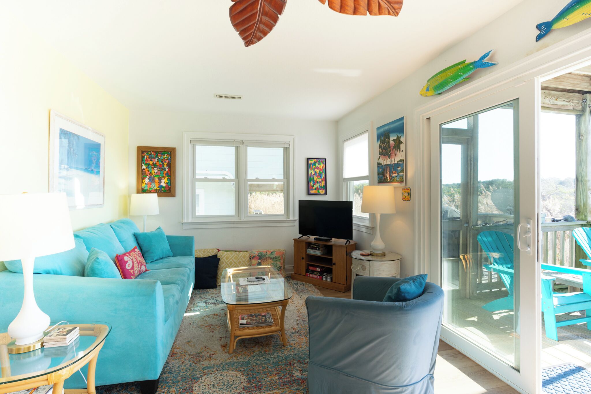 Un salon comprenant un canapé, une table basse en verre, un téléviseur et une vue sur une terrasse extérieure avec des chaises, accessible par une porte-fenêtre coulissante.
