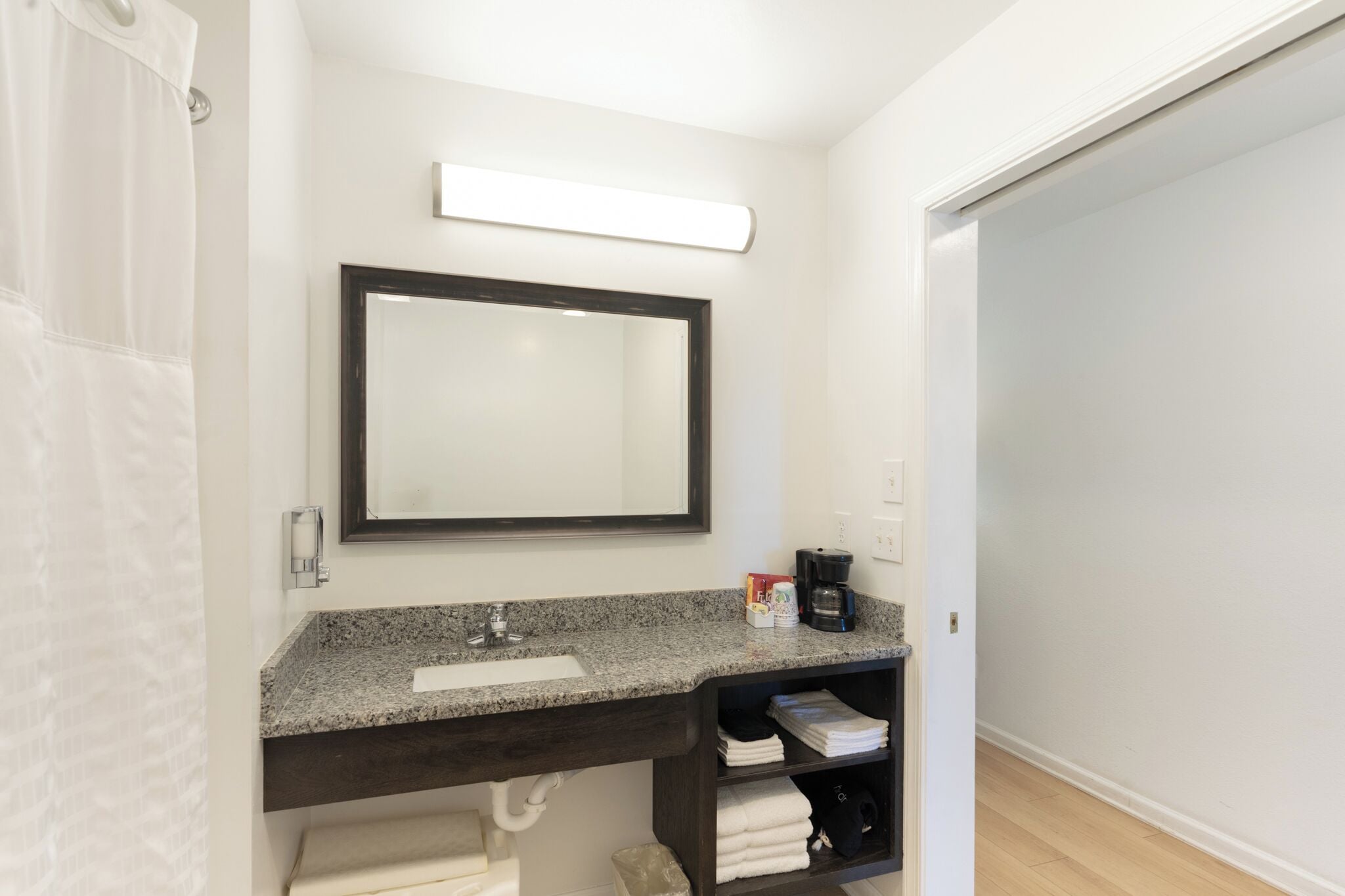 A bathroom with a granite countertop, a dark wooden vanity, a large mirror, a towel rack, and a coffee maker.