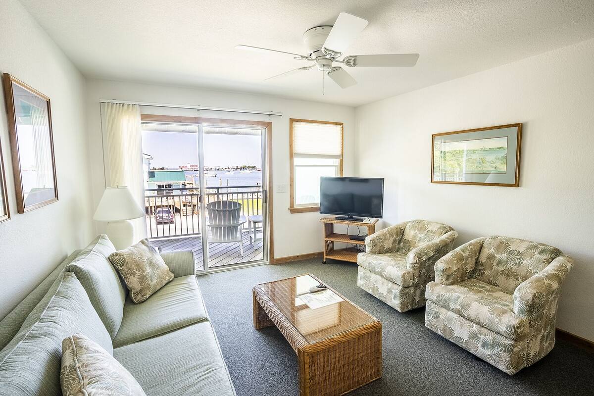 Ein Wohnzimmer mit Sofa, zwei Sesseln, einem Couchtisch, einem Fernseher auf einem Schrank und einem Balkon mit Blick auf das Wasser.