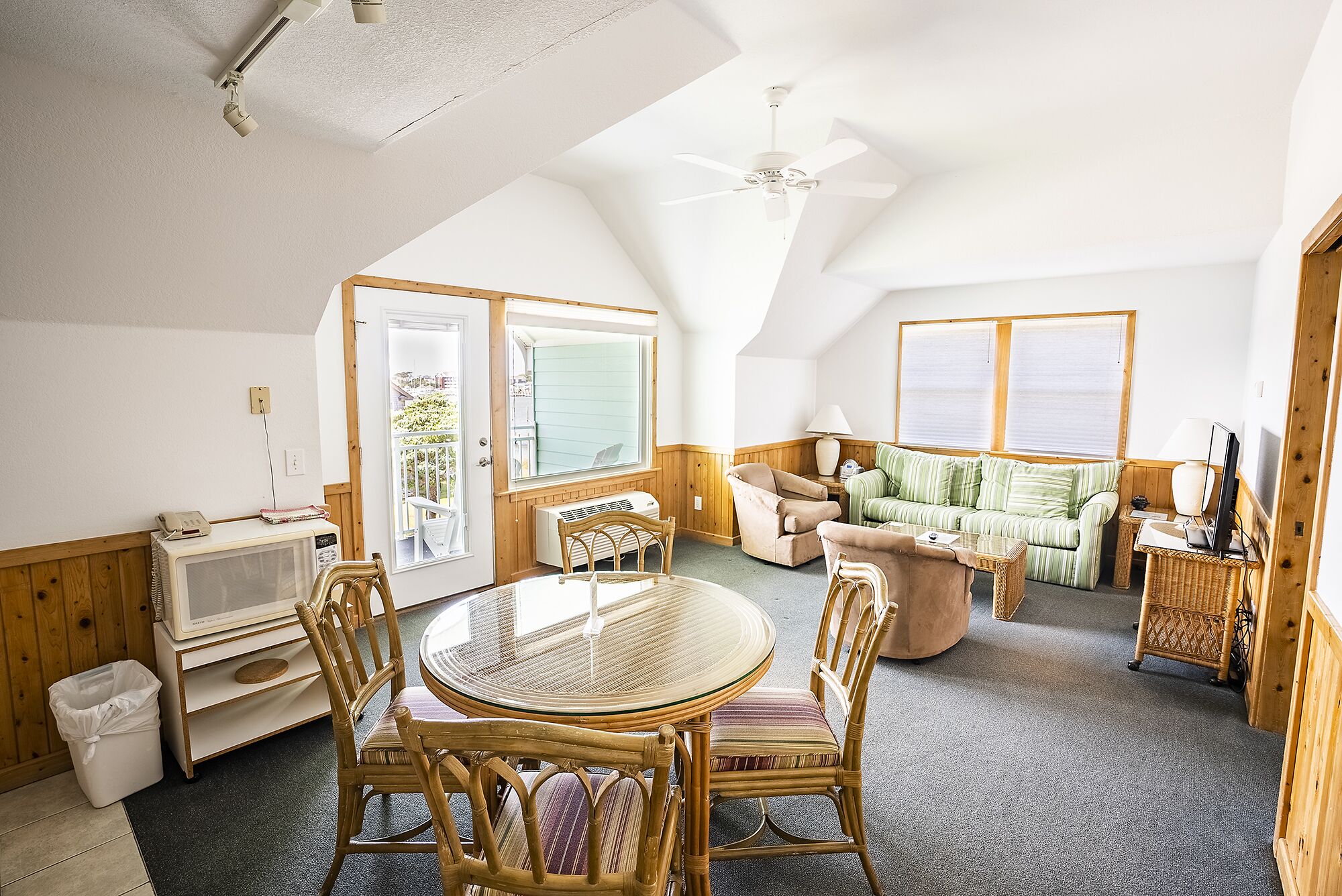 A cozy, well-lit room with a high ceiling, a ceiling fan, a round glass table with wicker chairs, a sofa, and a television.