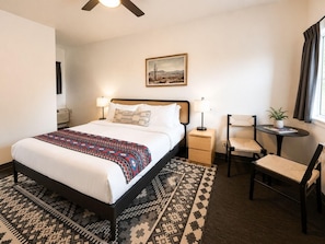 Une chambre d’hôtel comprenant un lit, des tables de chevet, une chaise, une petite table, un ventilateur de plafond, un tableau et une fenêtre avec des rideaux.