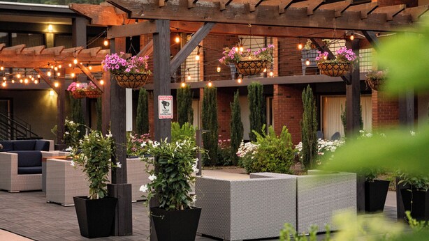An outdoor patio area with wicker furniture, hanging flower baskets, and a brick building in the background. - The Beaucatcher – A Boutique Motel (Asheville)