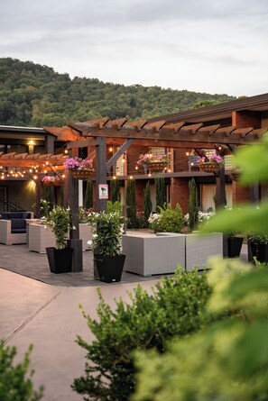 An outdoor patio area with wicker furniture, hanging flower baskets, and a brick building in the background. - The Beaucatcher – A Boutique Motel (Asheville)
