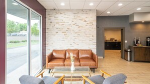 A reception area with a brown leather sofa, a glass coffee table, and two grey armchairs. A brick-patterned wall and a large window are visible. - The Beaucatcher – A Boutique Motel (Asheville)