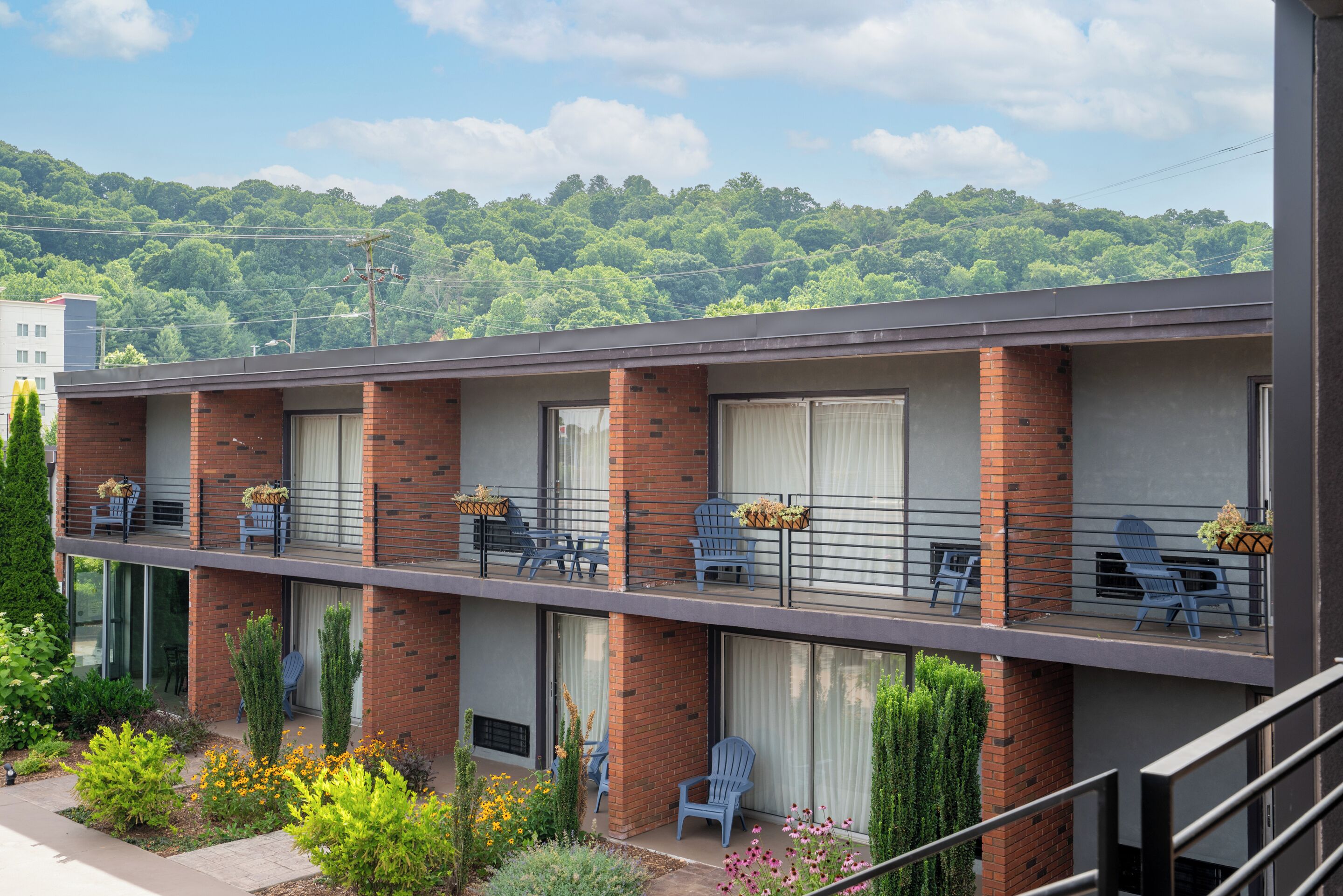 A two-story motel with brick exterior, balconies with blue chairs and potted plants, and a garden with various plants and flowers.