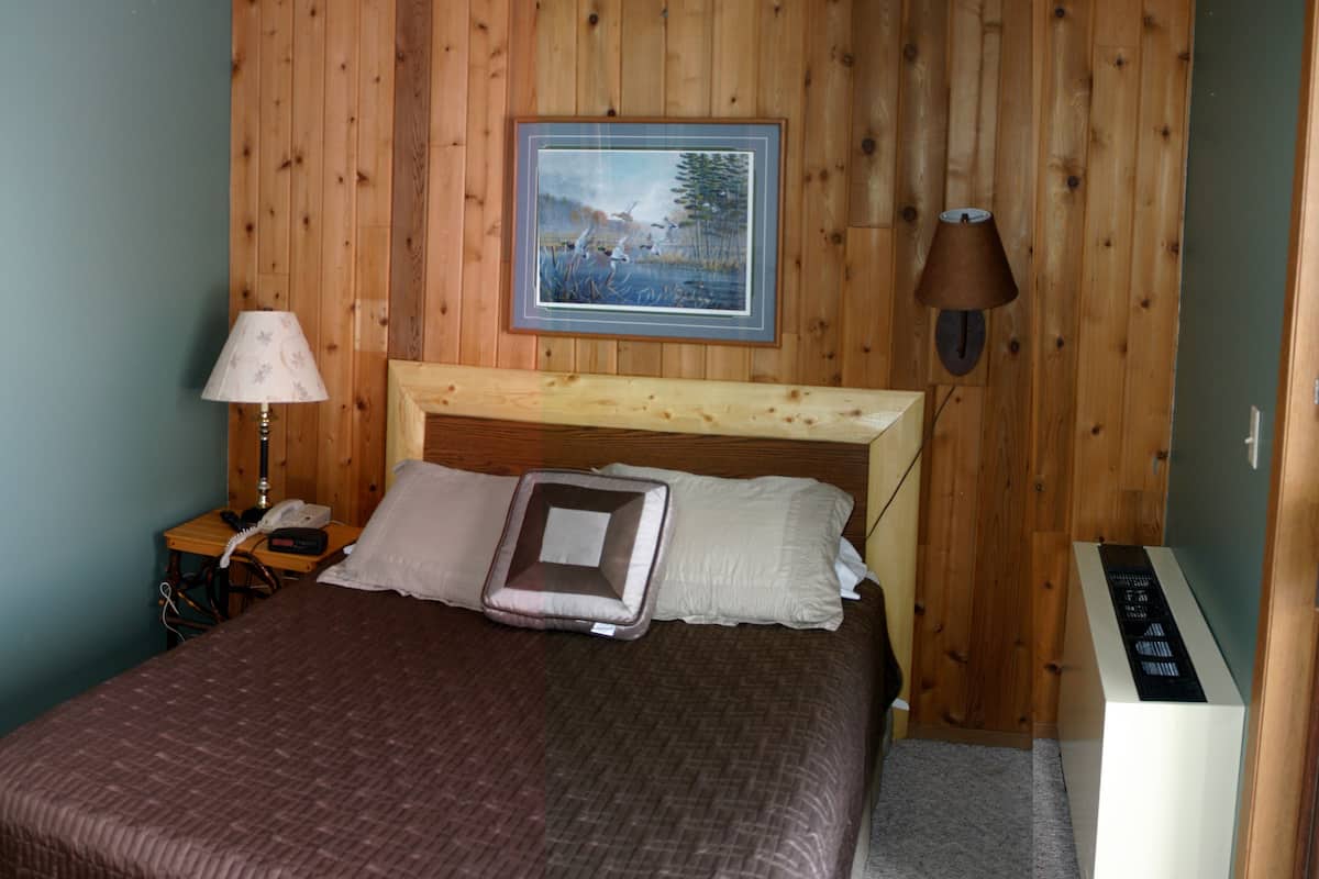 A bedroom with a wooden headboard, a framed picture on the wall, a bedside lamp, a telephone, and a thermostat.