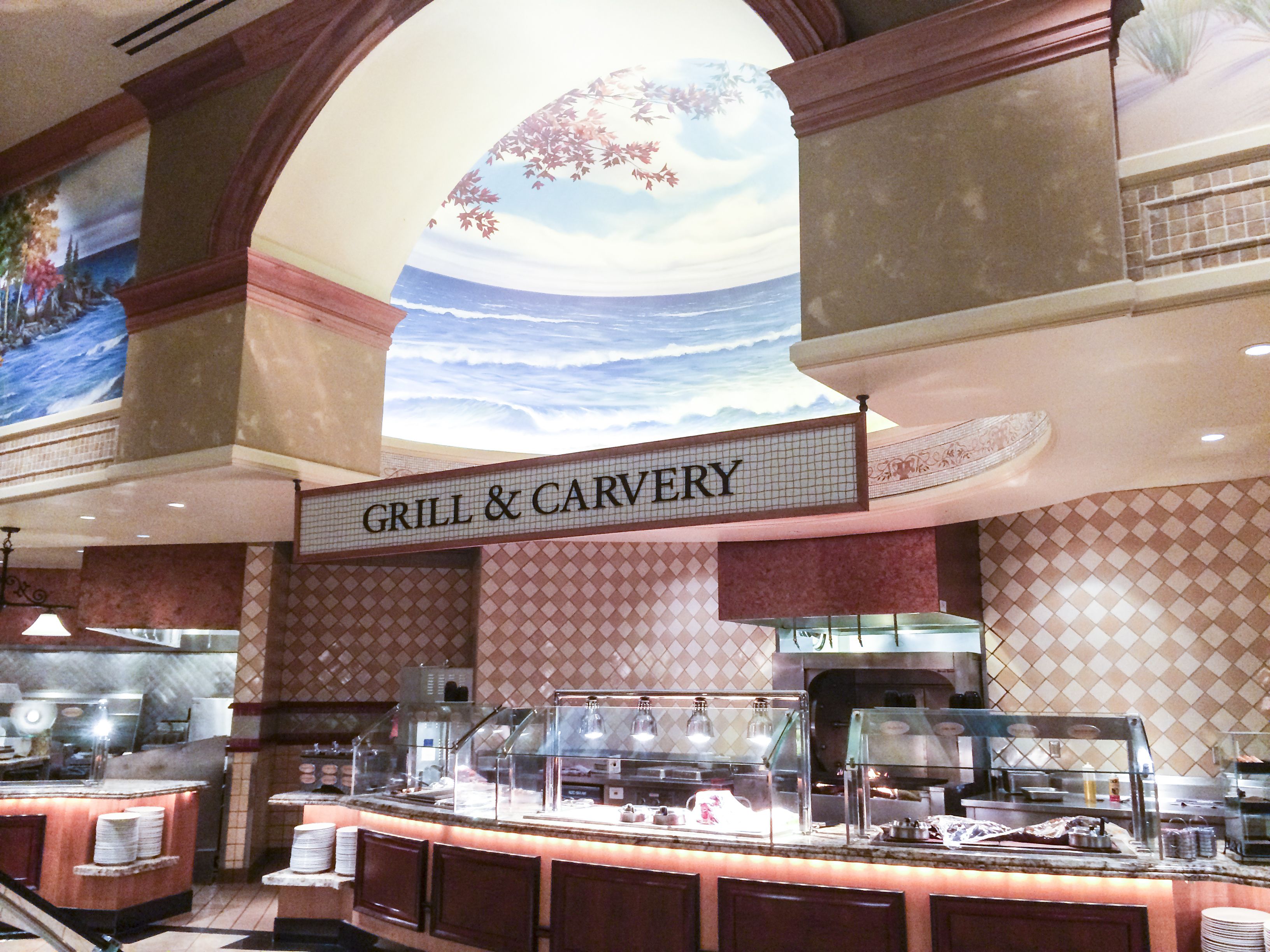 Ein Restaurantbereich mit einem Schild 'GRILL & CARVERY', einer Buffettheke mit Speisenauslagen und einem Wandgemälde, das eine Strandansicht zeigt.