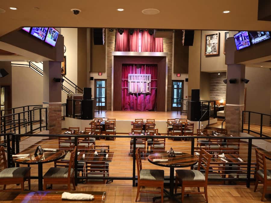 A restaurant with a stage and curtains in the center, wooden tables and chairs, and multiple televisions on the walls.