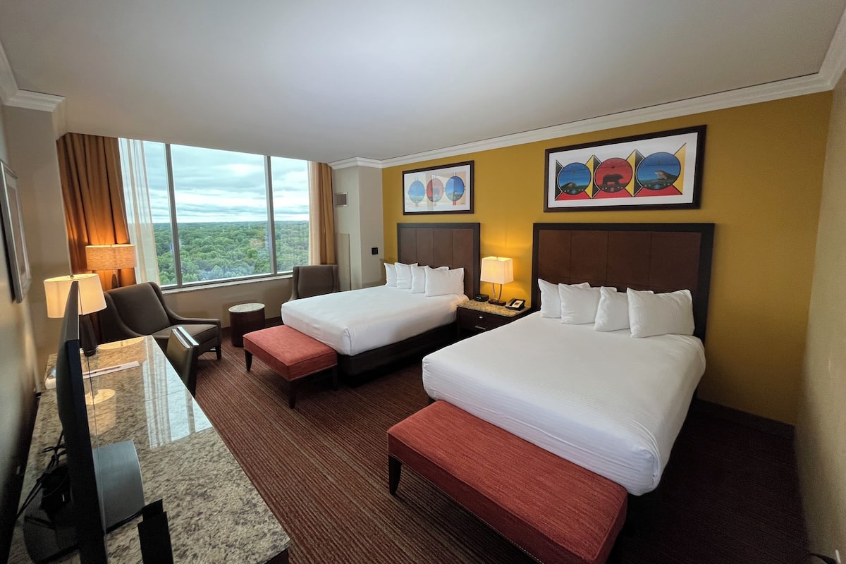 Ein Hotelzimmer mit zwei Betten, einem Schreibtisch, einem Stuhl, einem kleinen Tisch und Blick ins Freie.