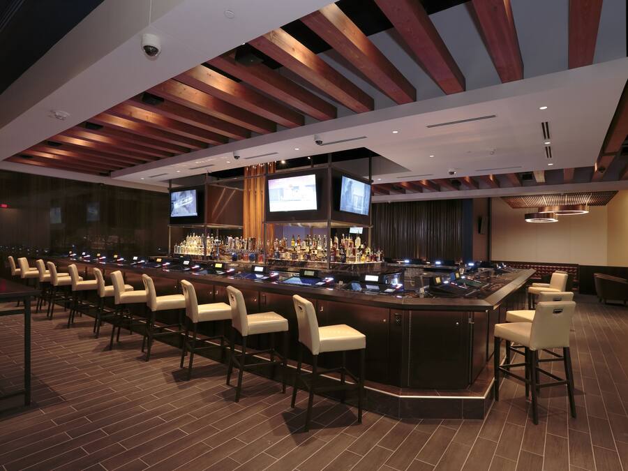 Uma área de bar com um balcão comprido, várias televisões e banquetas.