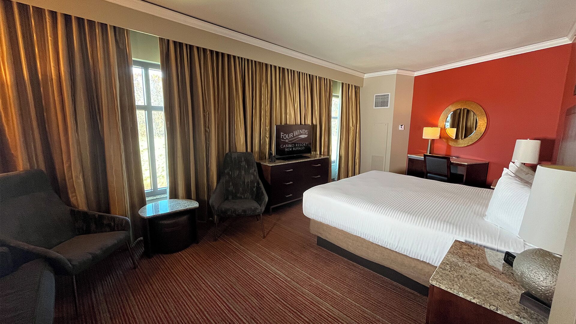 Quarto de hotel com uma cama grande, uma escrivaninha, uma cadeira, uma televisão e uma janela com cortinas.