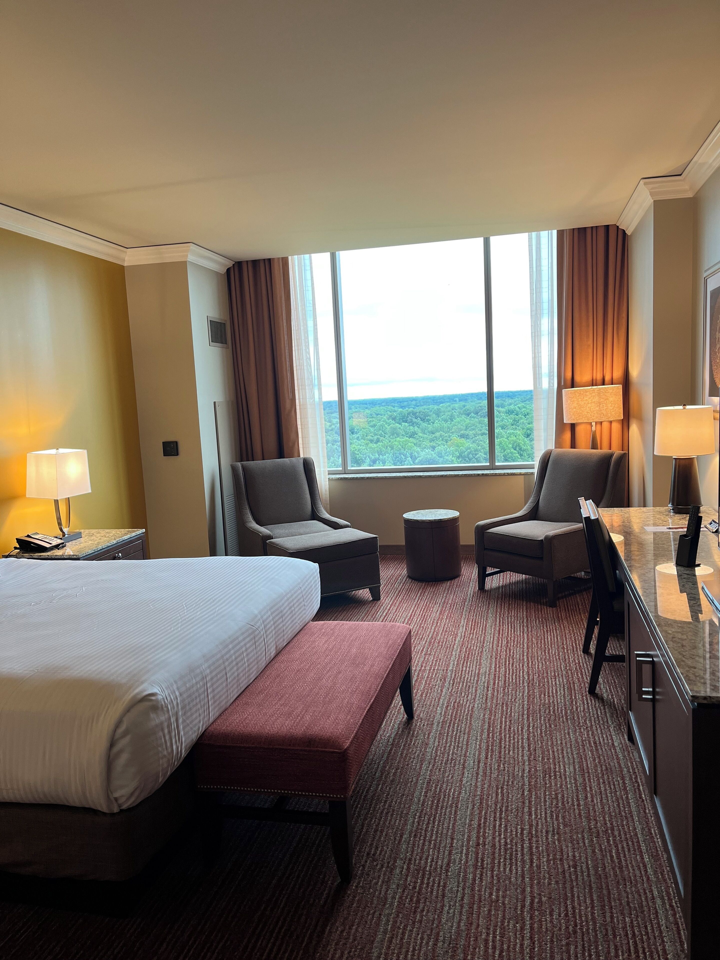 Habitación de hotel con una cama grande, dos sillones, un pequeño reposapiés, un escritorio con lámpara y vistas al exterior.