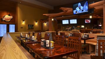 Un restaurante con mesas y sillas de madera, una zona de bar y una pantalla de televisión mostrando un partido de hockey.