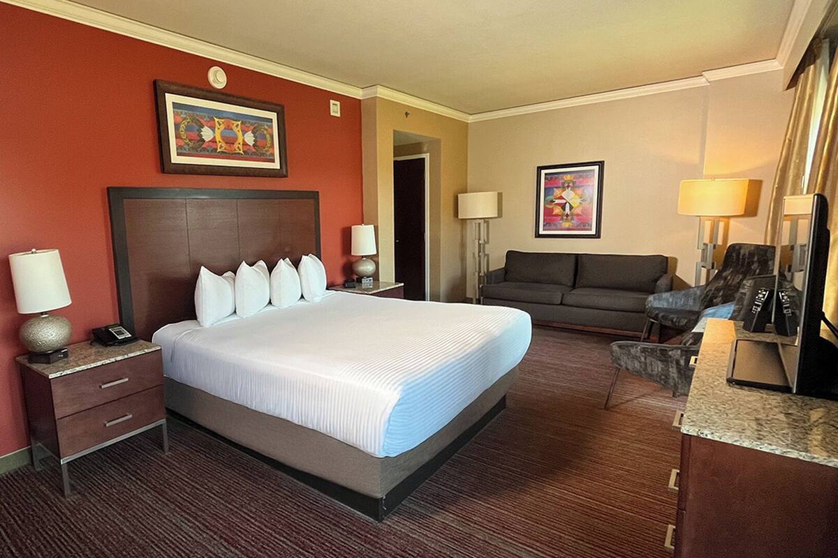 Ein Hotelzimmer mit einem großen Bett, einem Sofa, einem Schreibtisch und einem Stuhl. Auf dem Schreibtisch stehen zwei Lampen und an der Wand hängt ein Bild.