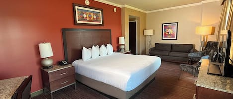 Una camera d'albergo con un letto grande, un divano, una scrivania e una sedia. Ci sono due lampade sulla scrivania e un quadro appeso al muro.