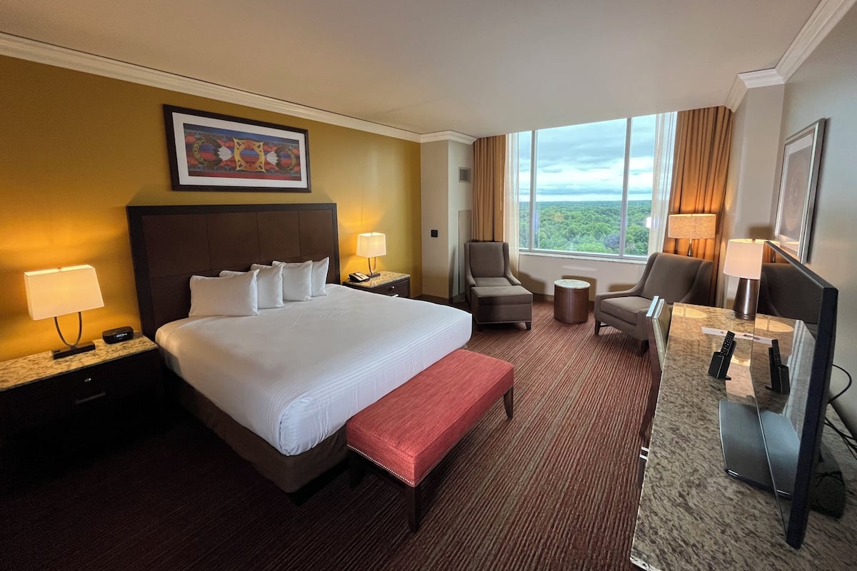 Ein Hotelzimmer mit einem großen Bett, zwei Nachttischen mit Lampen, einem Schreibtisch mit Stuhl, einem Fernseher und einem Fenster mit Blick ins Freie.