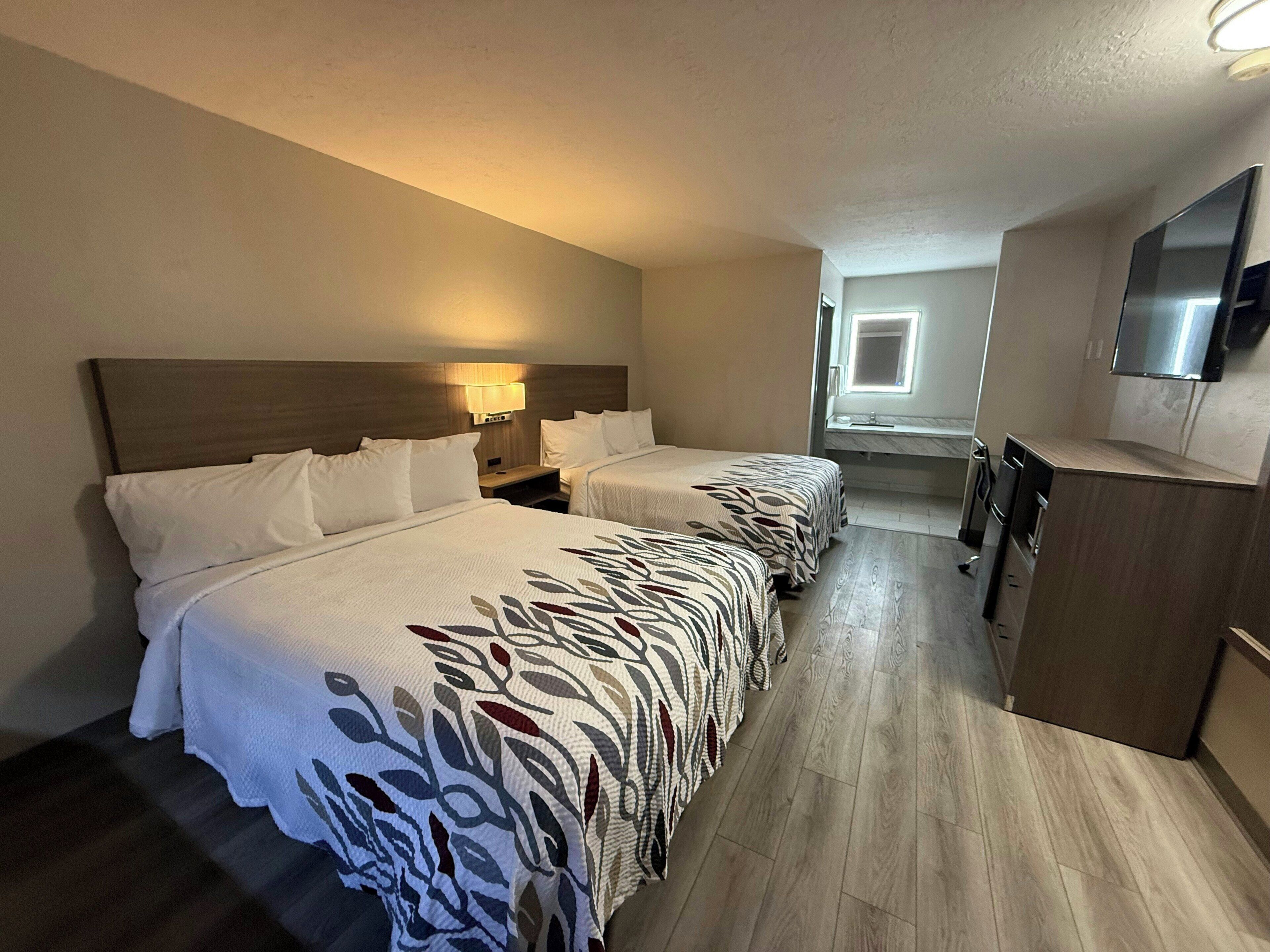 A hotel room with two beds, a TV, a desk, and a bathroom visible through an open door.