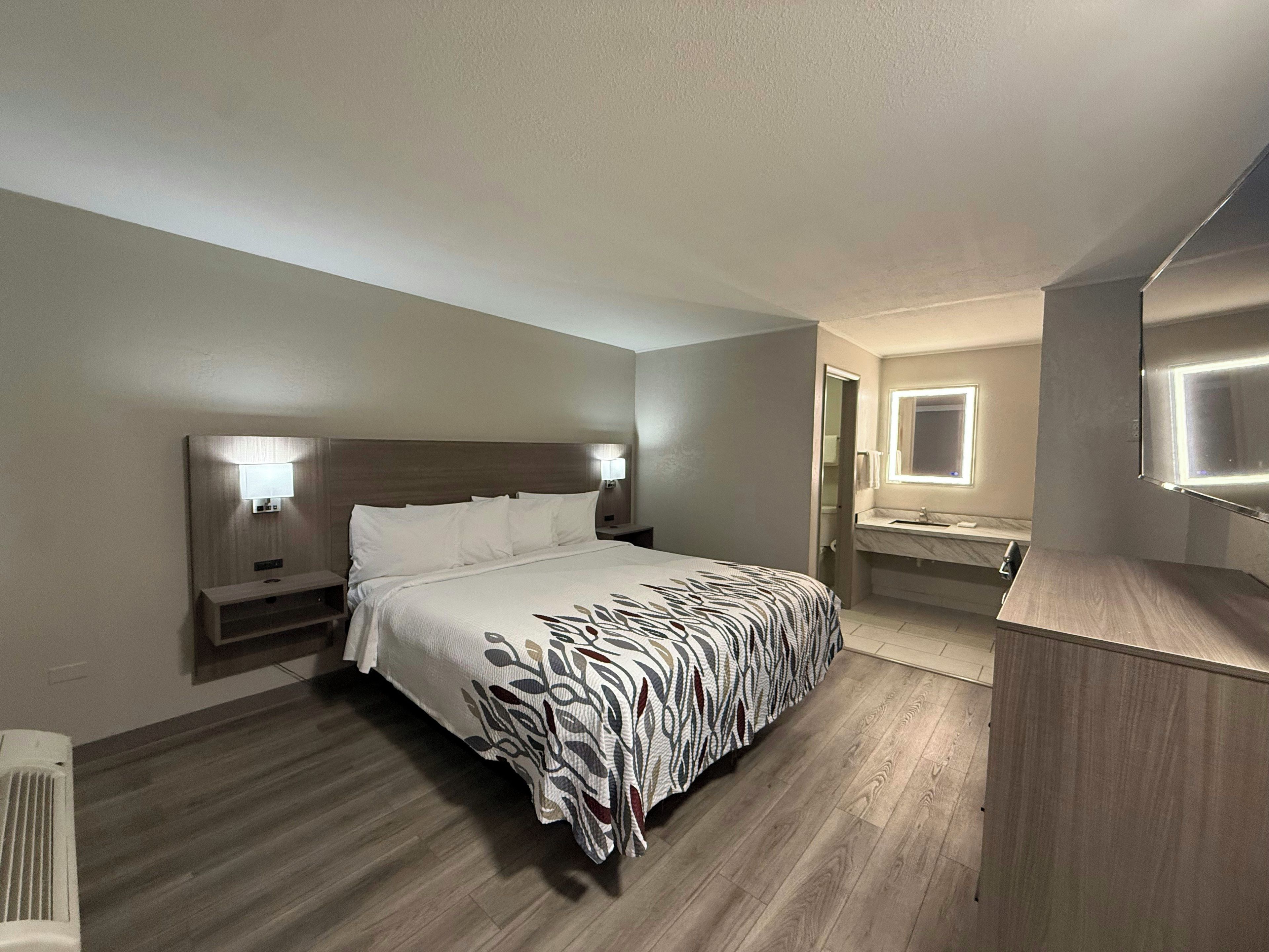 A hotel room with a bed, bedside lamps, a bathroom with a mirror and sink, and a wooden floor.