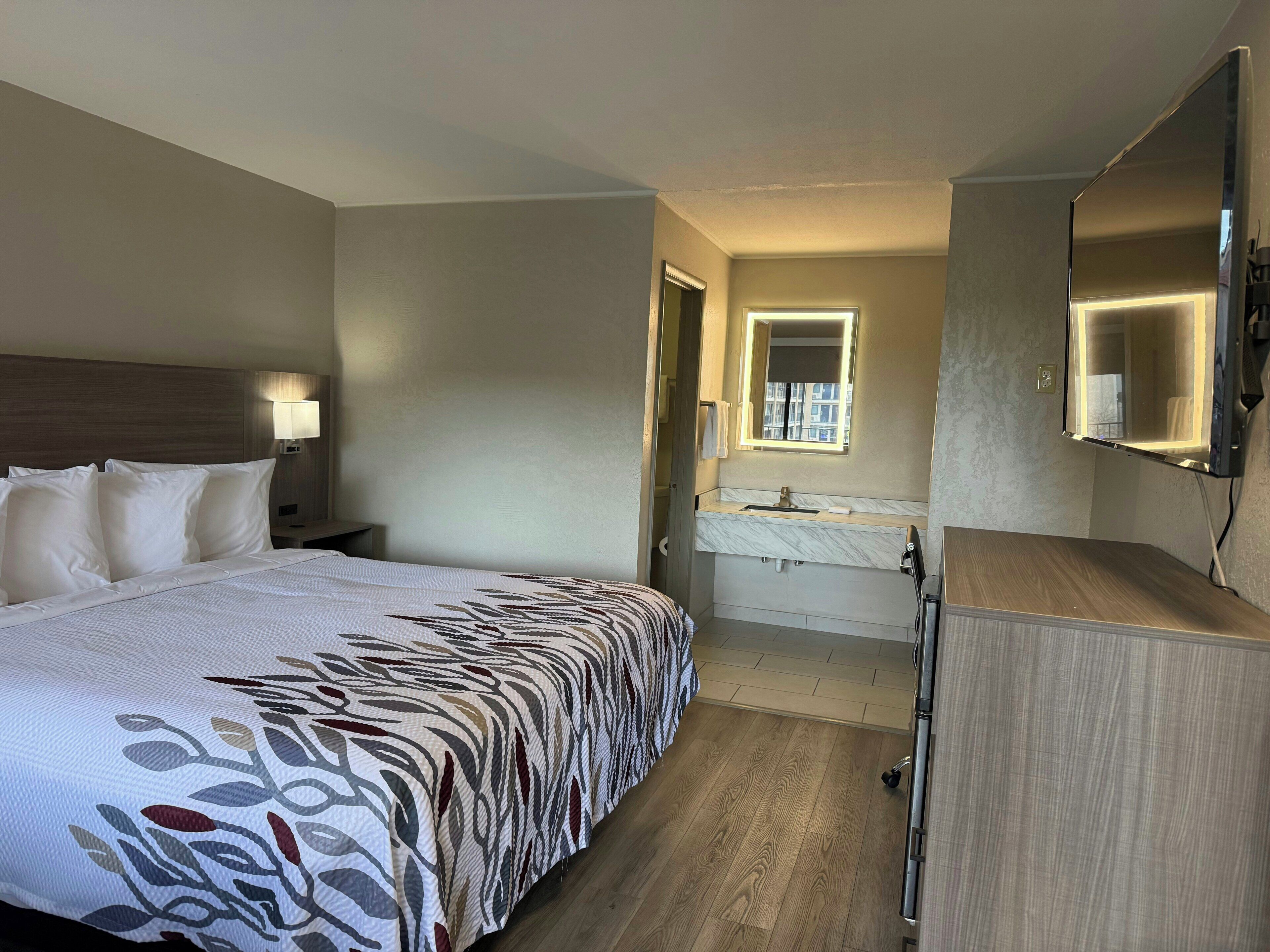 A hotel room with a bed, bedside lamp, a bathroom with a mirror, sink, and a wooden cabinet.