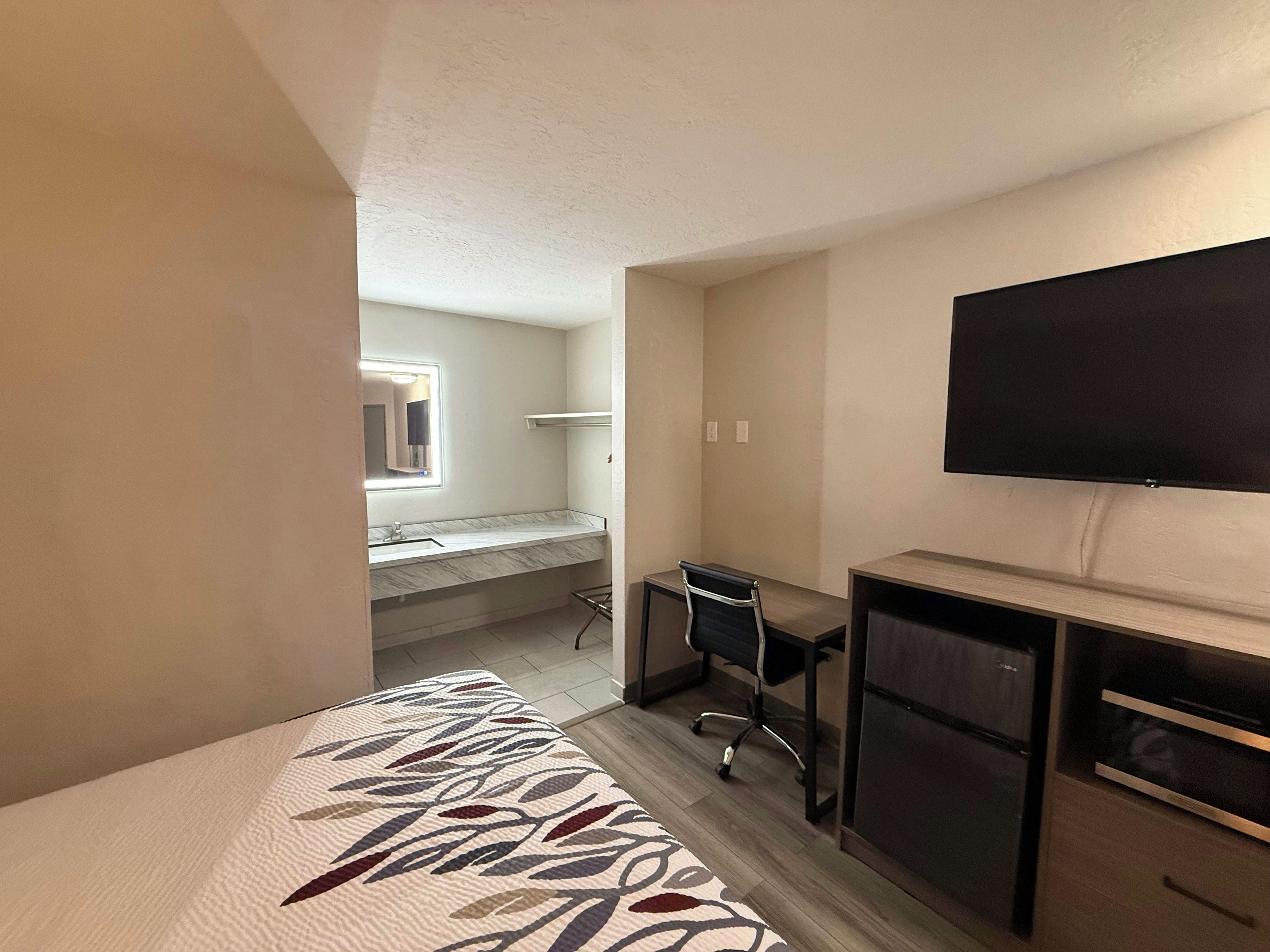 A hotel room with a bed, a desk, a chair, a television, and a bathroom with a mirror and sink.