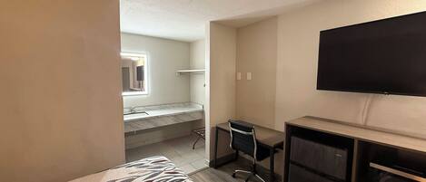 A hotel room with a bed, a desk, a chair, a television, and a bathroom with a mirror and sink.