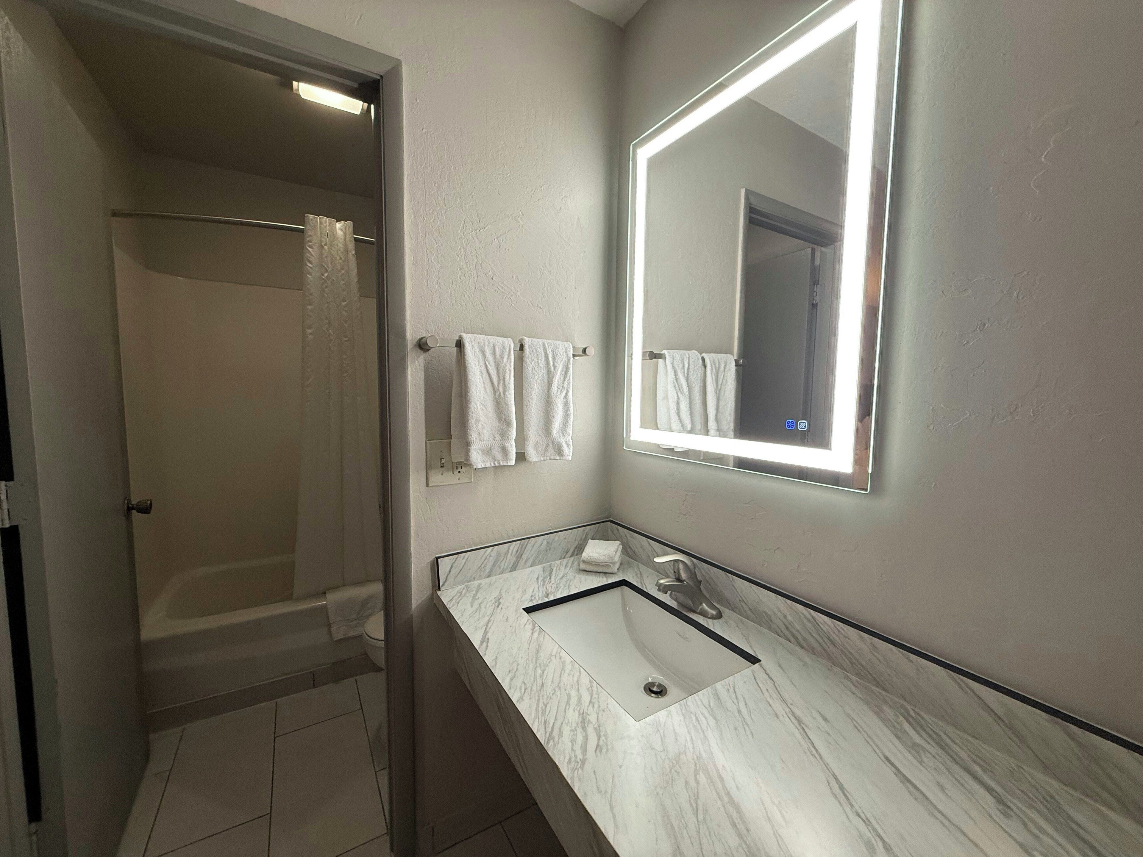 A bathroom with a shower, a sink with a marble countertop, and a mirror with a light.