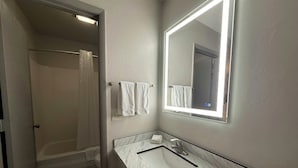 A bathroom with a shower, a sink with a marble countertop, and a mirror with a light. - Red Roof Reno (Reno)