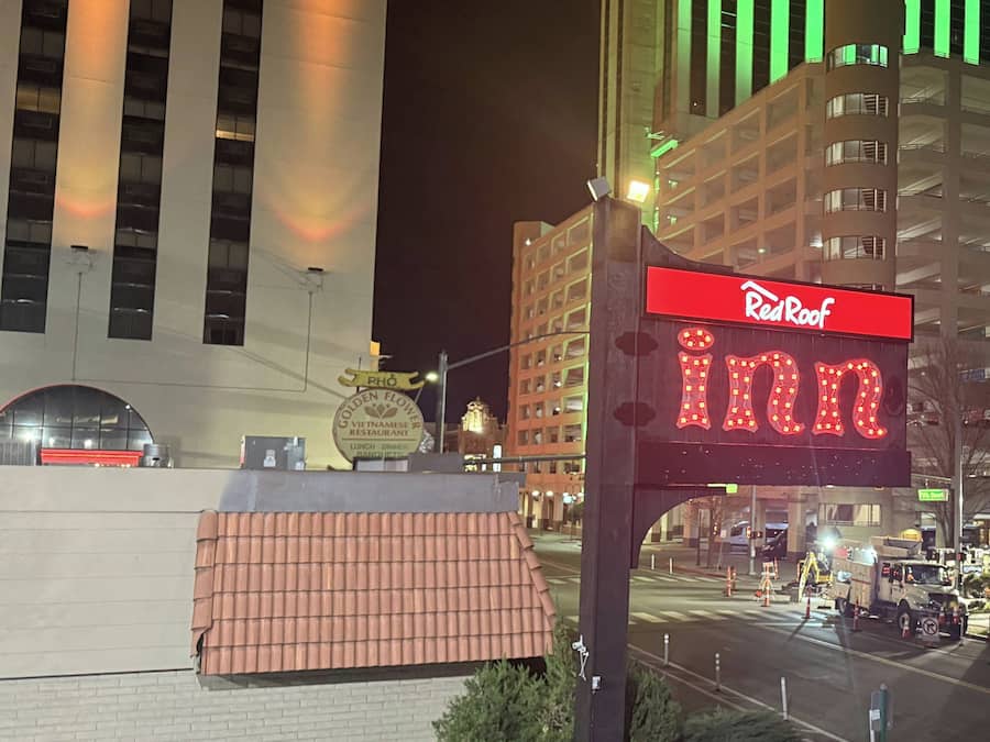Señal de Red Roof Inn con una marquesina de neón, una pared de ladrillo y un edificio con luces verdes verticales al fondo.