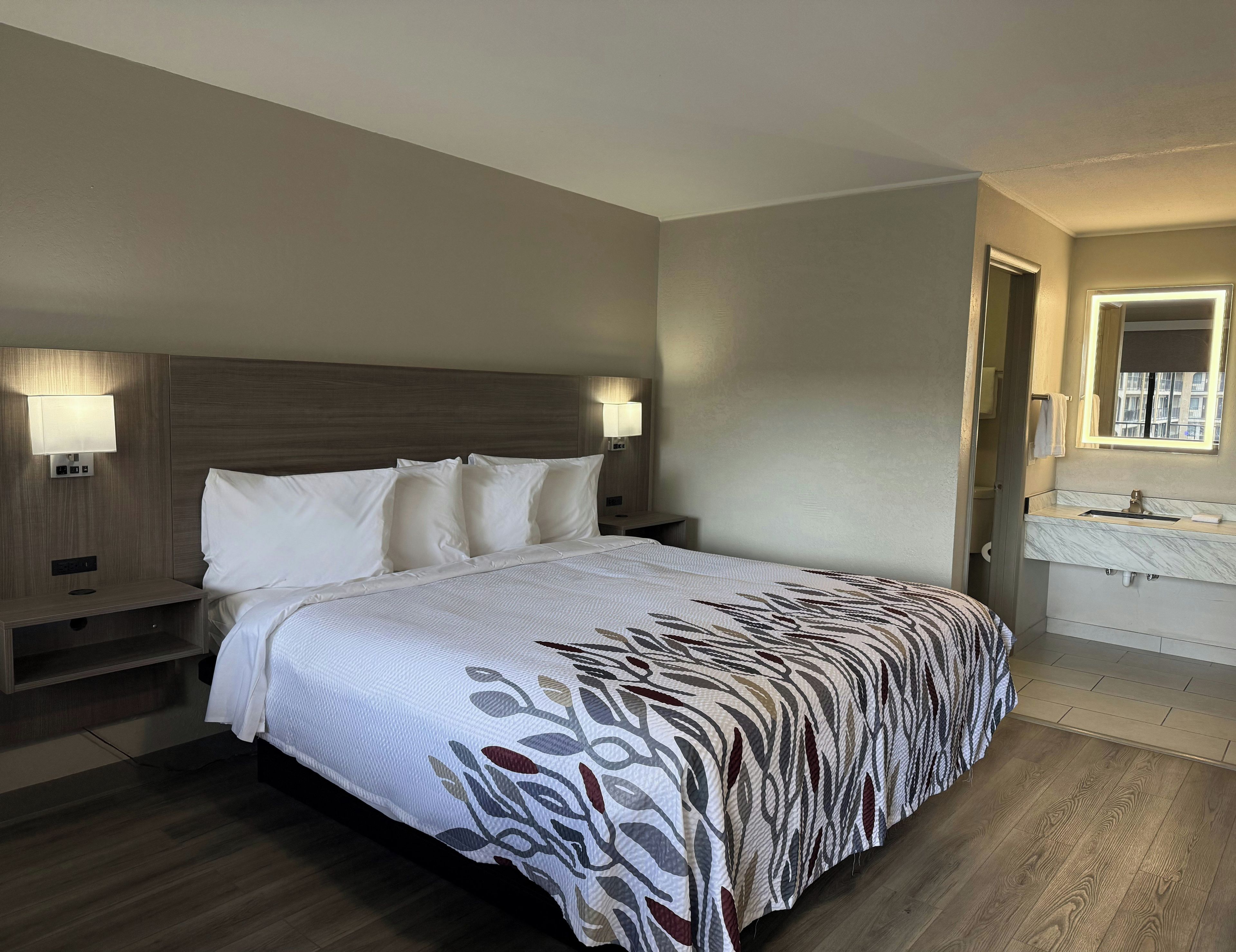 A hotel room with a large bed, bedside lamps, a nightstand, a bathroom with a mirror and sink, and a wooden floor.
