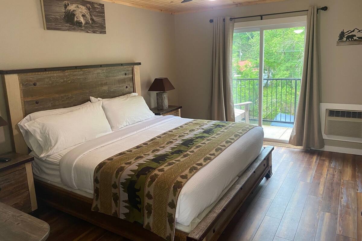 Ein Schlafzimmer mit einem hölzernen Bett, einem Nachttisch, einer Lampe, einem Deckenventilator und Blick ins Freie durch eine Schiebetür.