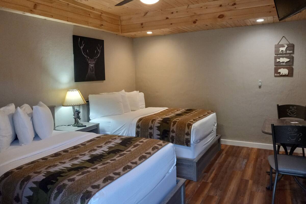 Ein Hotelzimmer mit zwei Betten, einem Deckenventilator, einer Lampe und einem an der Wand befestigten Schild, das Jagd-, Fischerei- und Laufsportarten anzeigt.